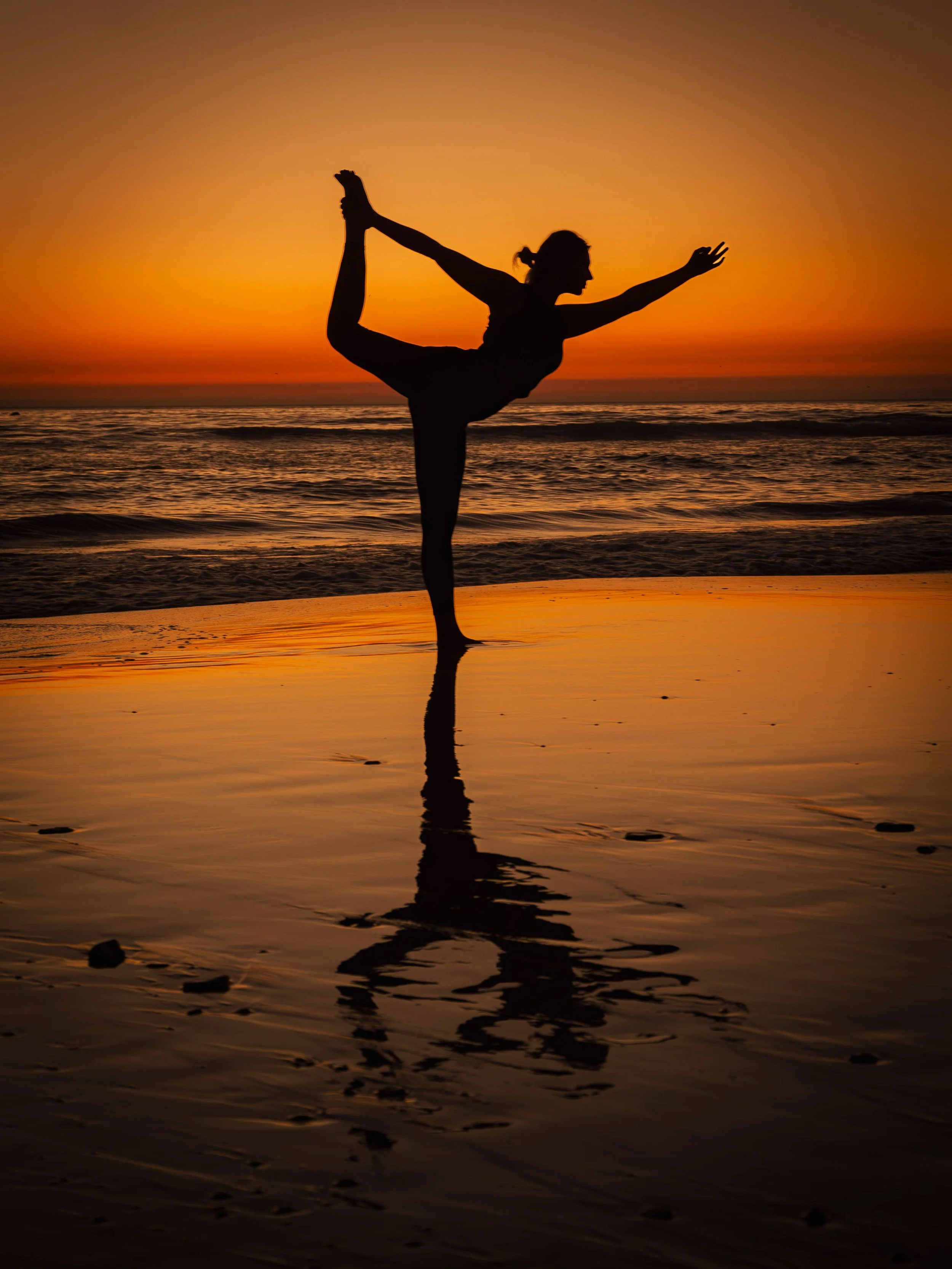Silhouette of a woman doing a yoga pose on the beach at sunset, reflected in the water.