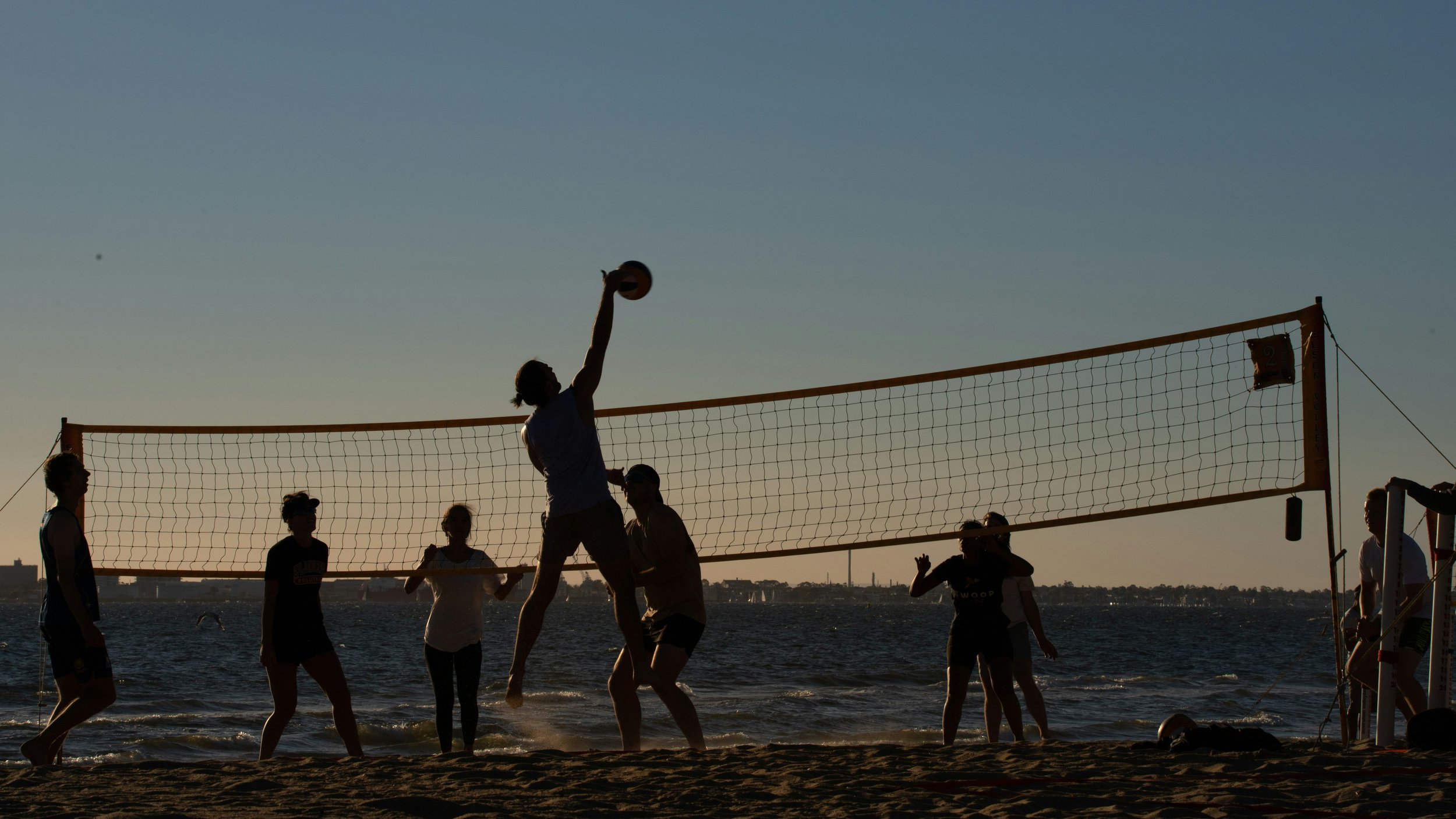 Silhouettes of people playing beach volleyball by the water during sunset.