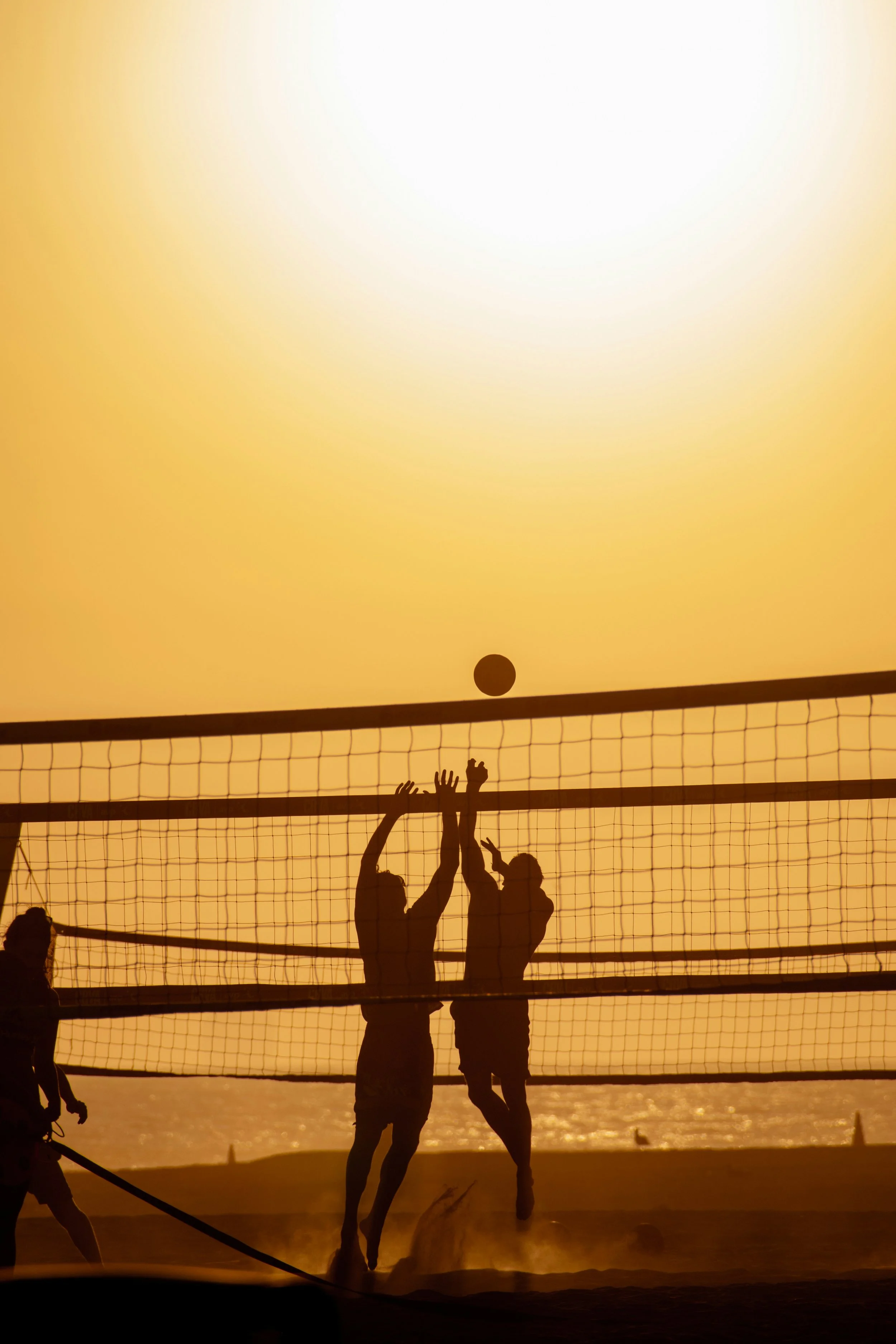 Silhouettes of people playing beach volleyball during sunset, with a glowing yellow sky and the ocean in the background.