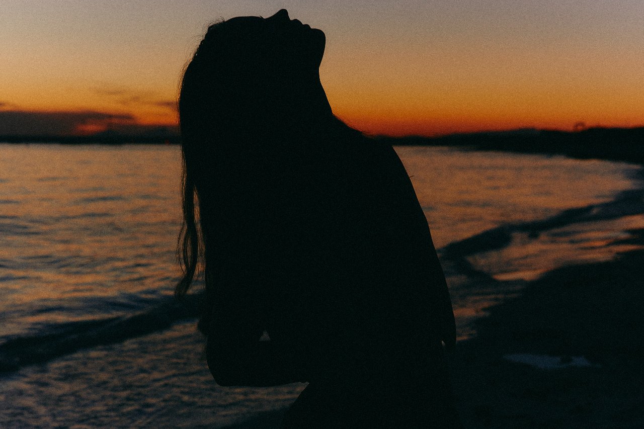 Silhouette of a person sitting by the water during sunset, with orange and purple sky in the background.