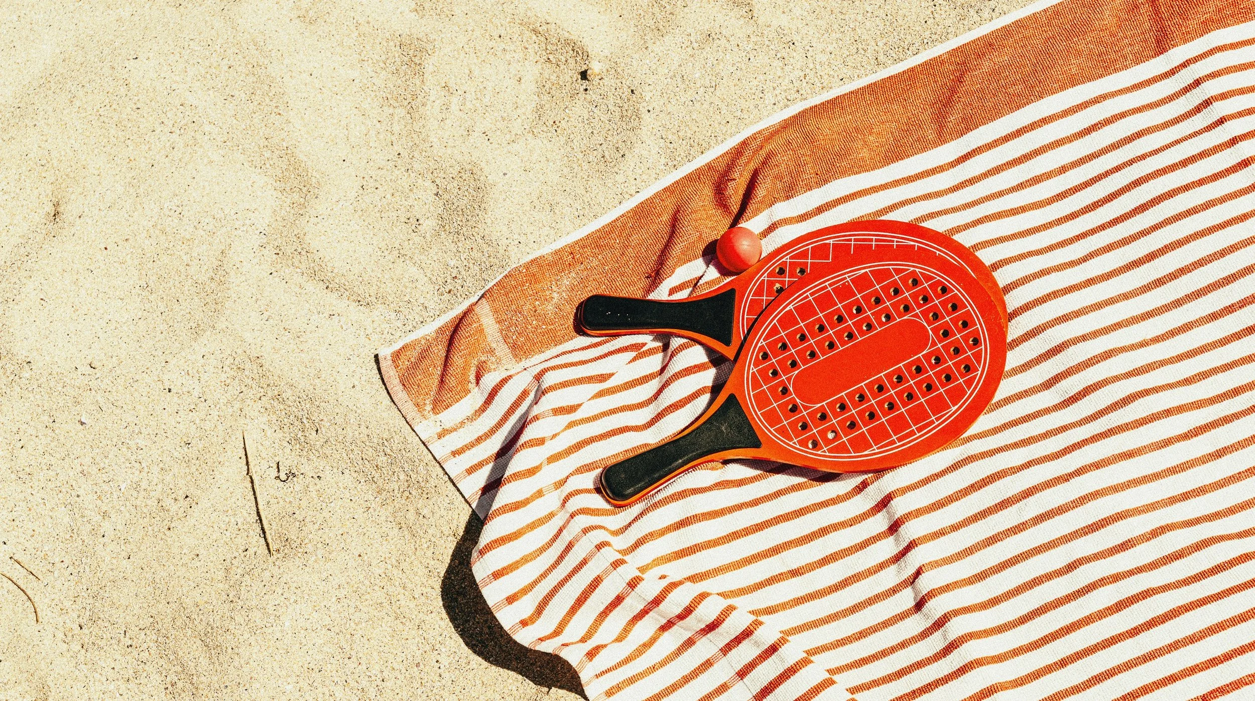 A red paddle and ball sit on an orange and white striped towel on sandy beach sand.