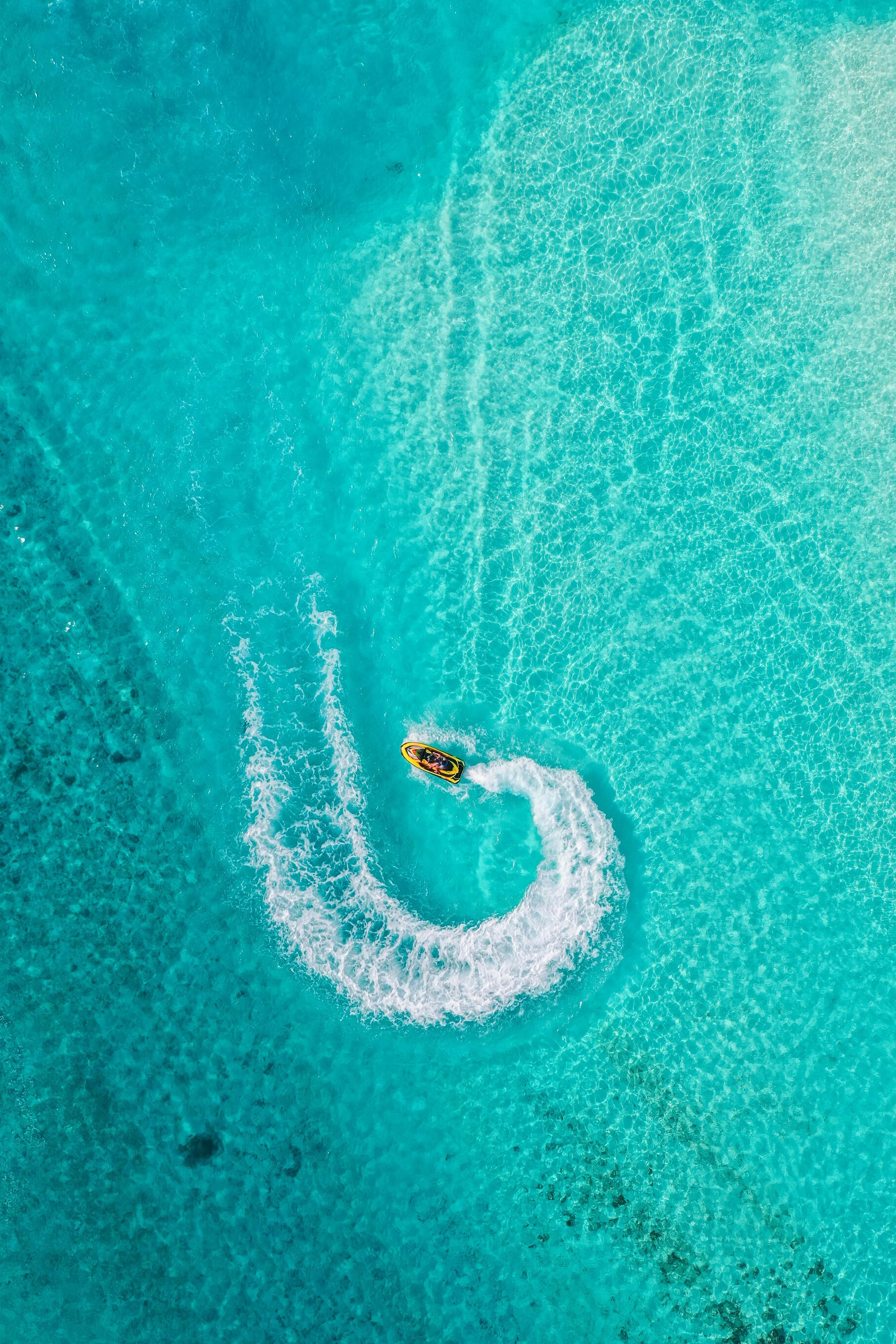 An aerial view of a boat speeding through clear, turquoise ocean water, creating a white foamy wake behind it.
