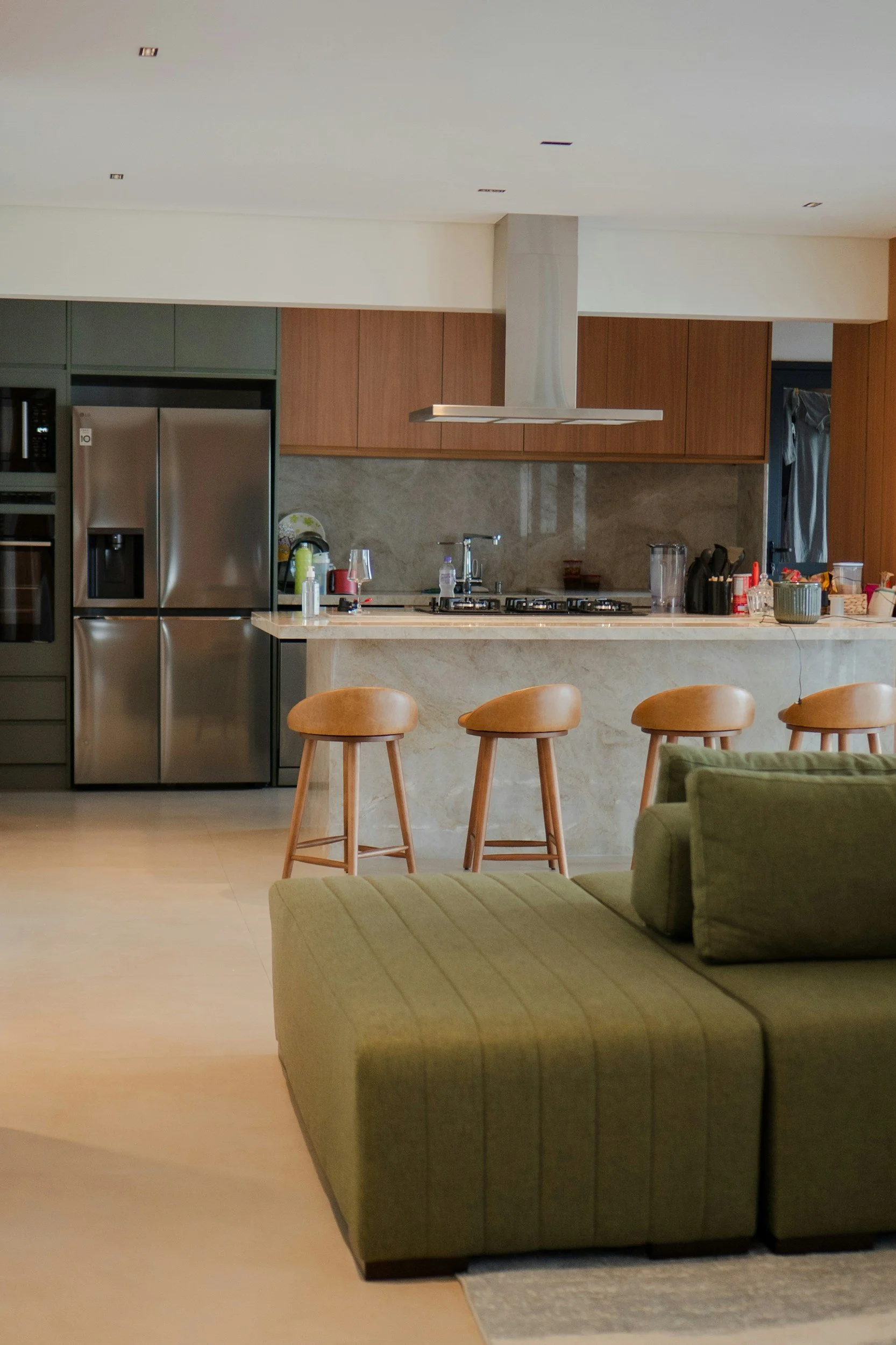 A modern kitchen with wooden cabinets, a stainless steel refrigerator, a gas stove, a kitchen island with barstools, and a green sofa in the foreground.