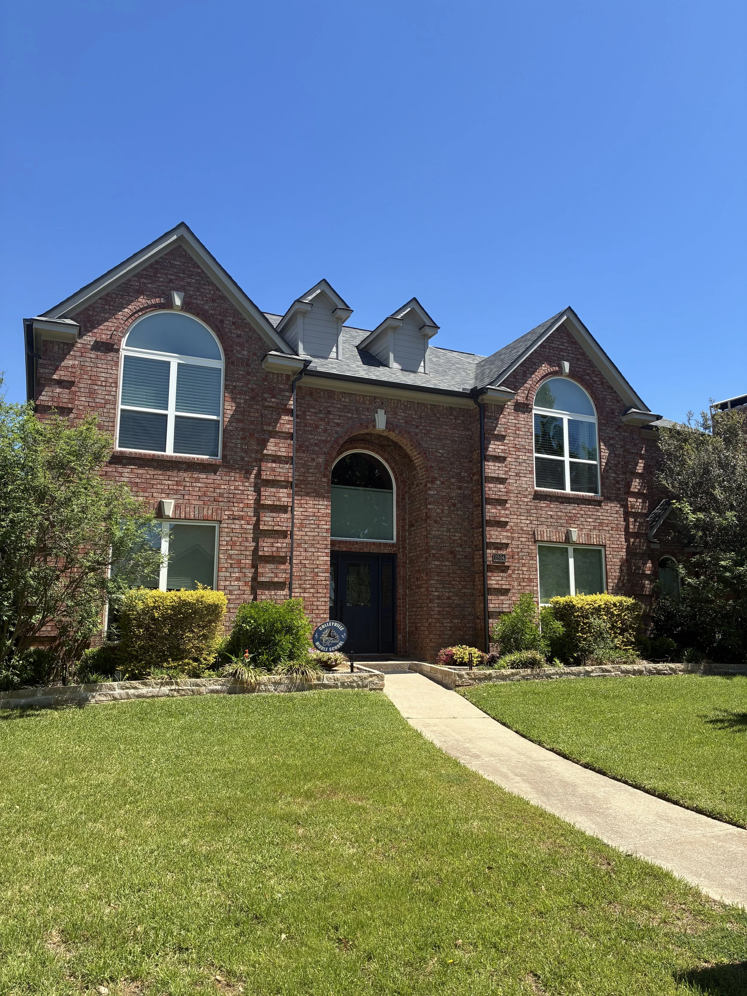 A two-story brick house with large arched windows, a gray slate roof, and a front yard with a walkway, green lawn, and shrubs under a clear blue sky.