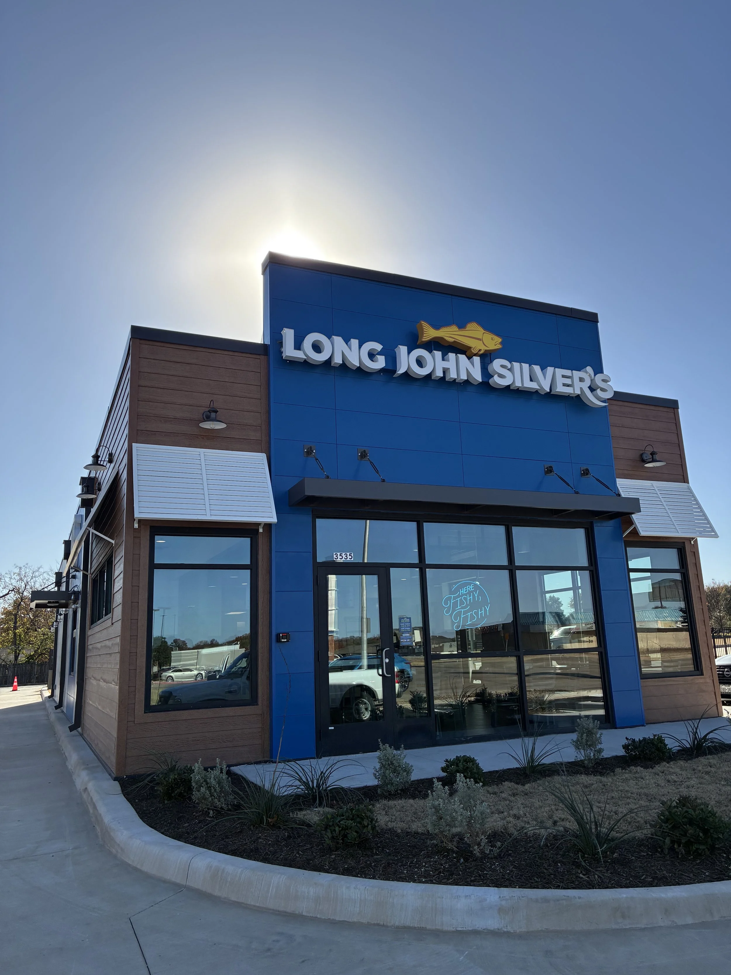 A storefront with a blue facade and brown wooden accents, displaying the name 'Long John Silver's' and a fish logo. The entrance has large glass doors, and there is a small landscaping area with plants in front.