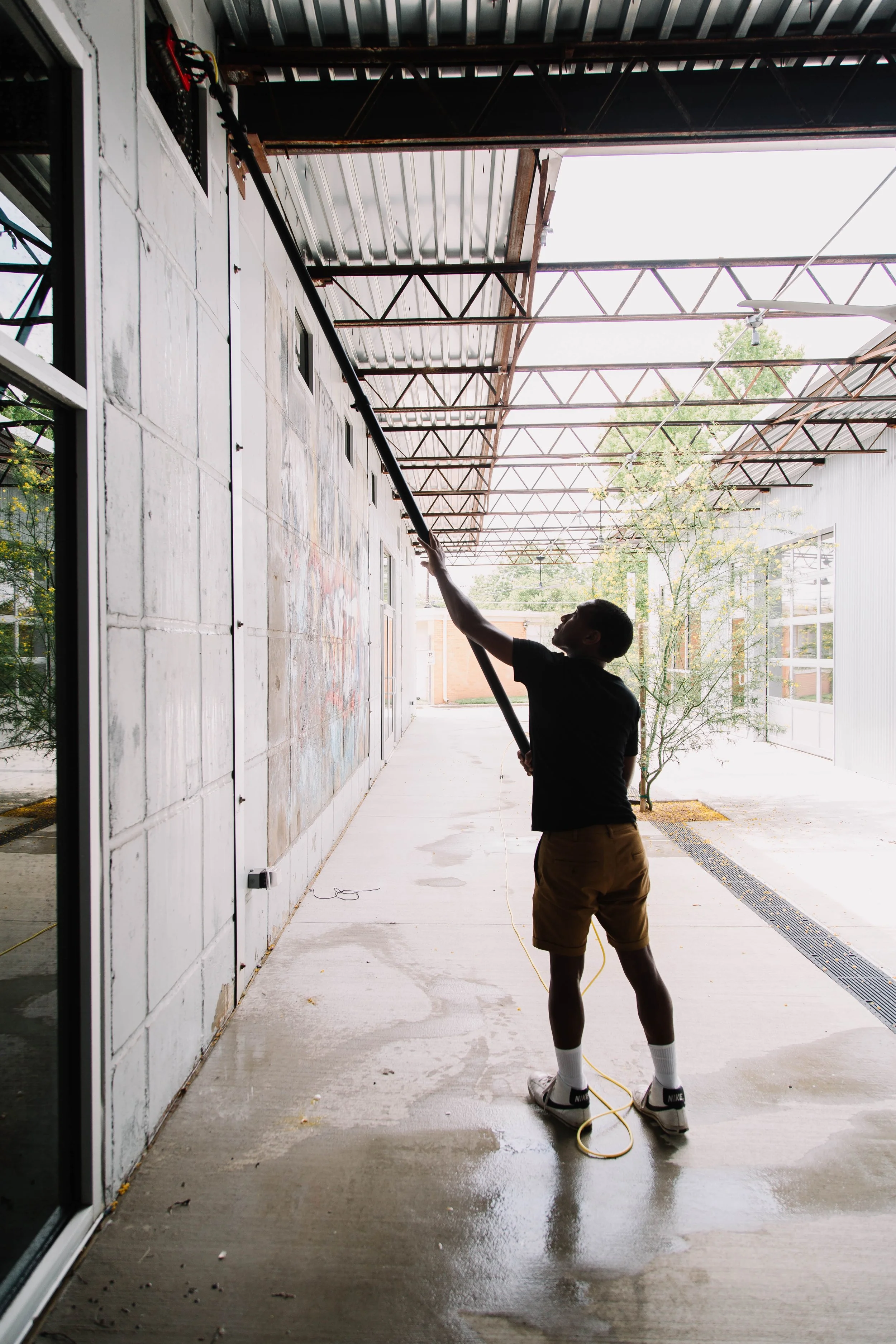 A person is pressure washing an outdoor concrete walkway under a metal roof structure, with trees and a building visible in the background.