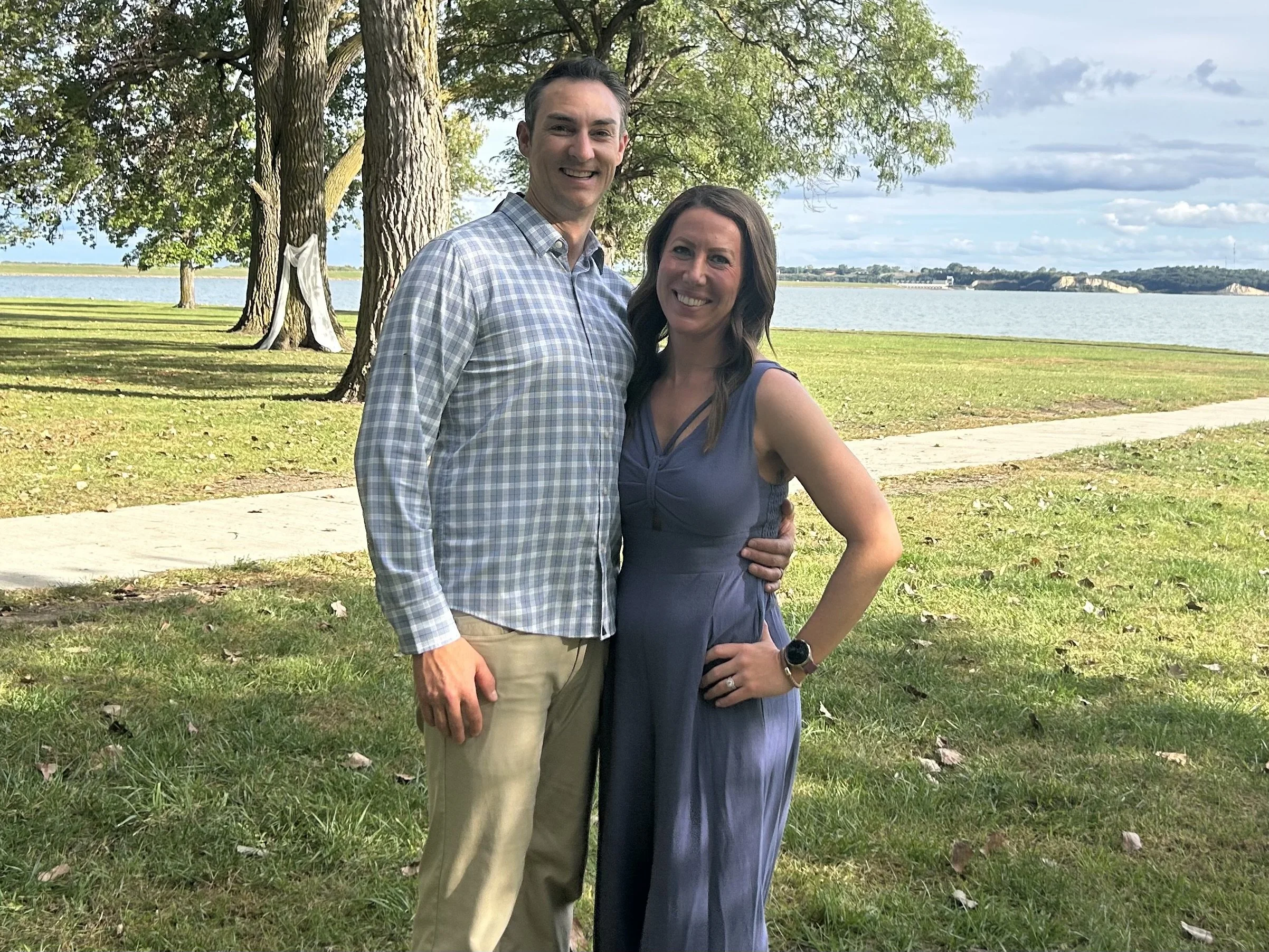 A smiling man and woman standing outdoors near a lake with trees in the background on a partly cloudy day.
