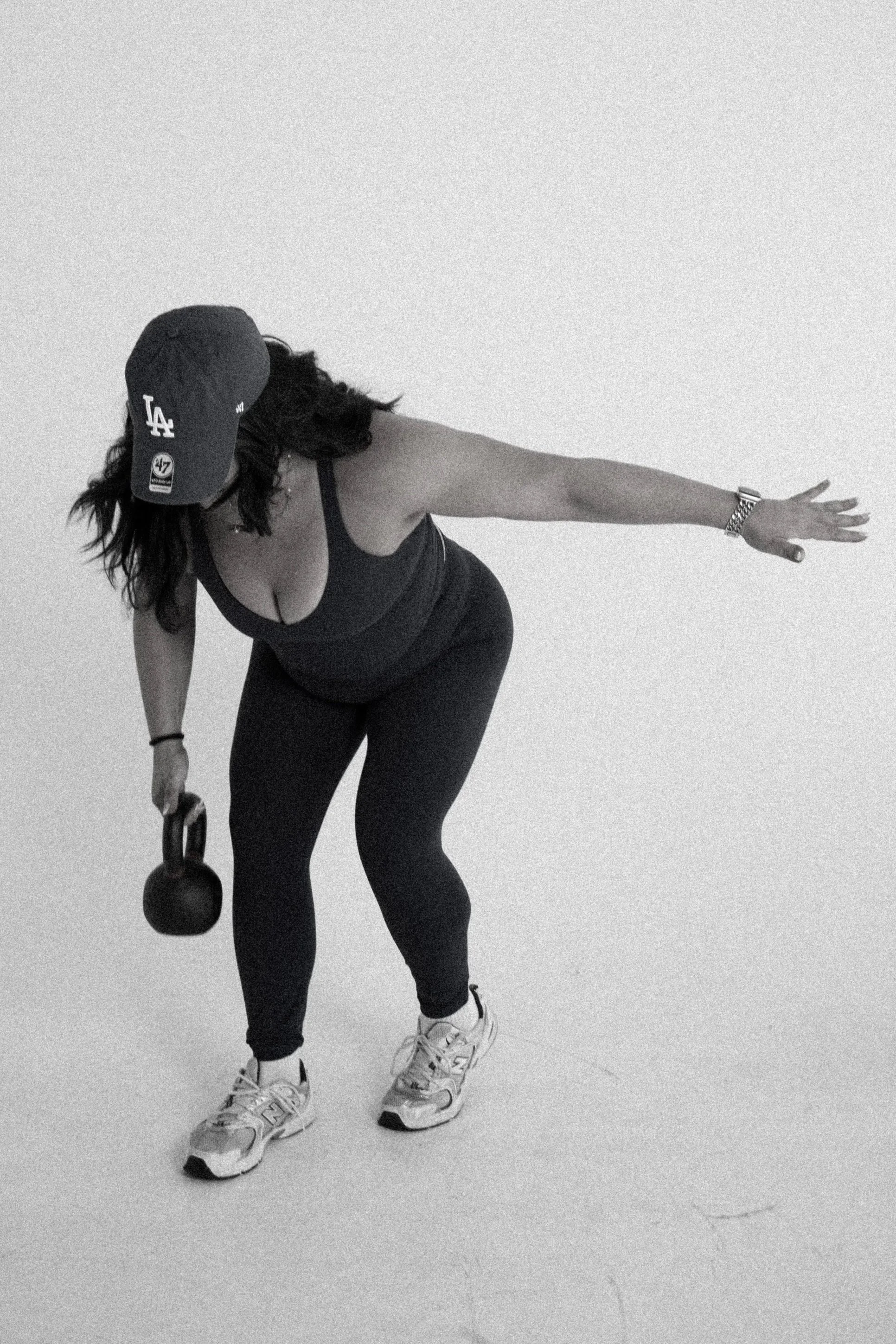 A woman in athletic wear and sneakers performing a kettlebell exercise, wearing a Los Angeles Dodgers baseball cap, standing on a plain light-colored floor against a plain wall.