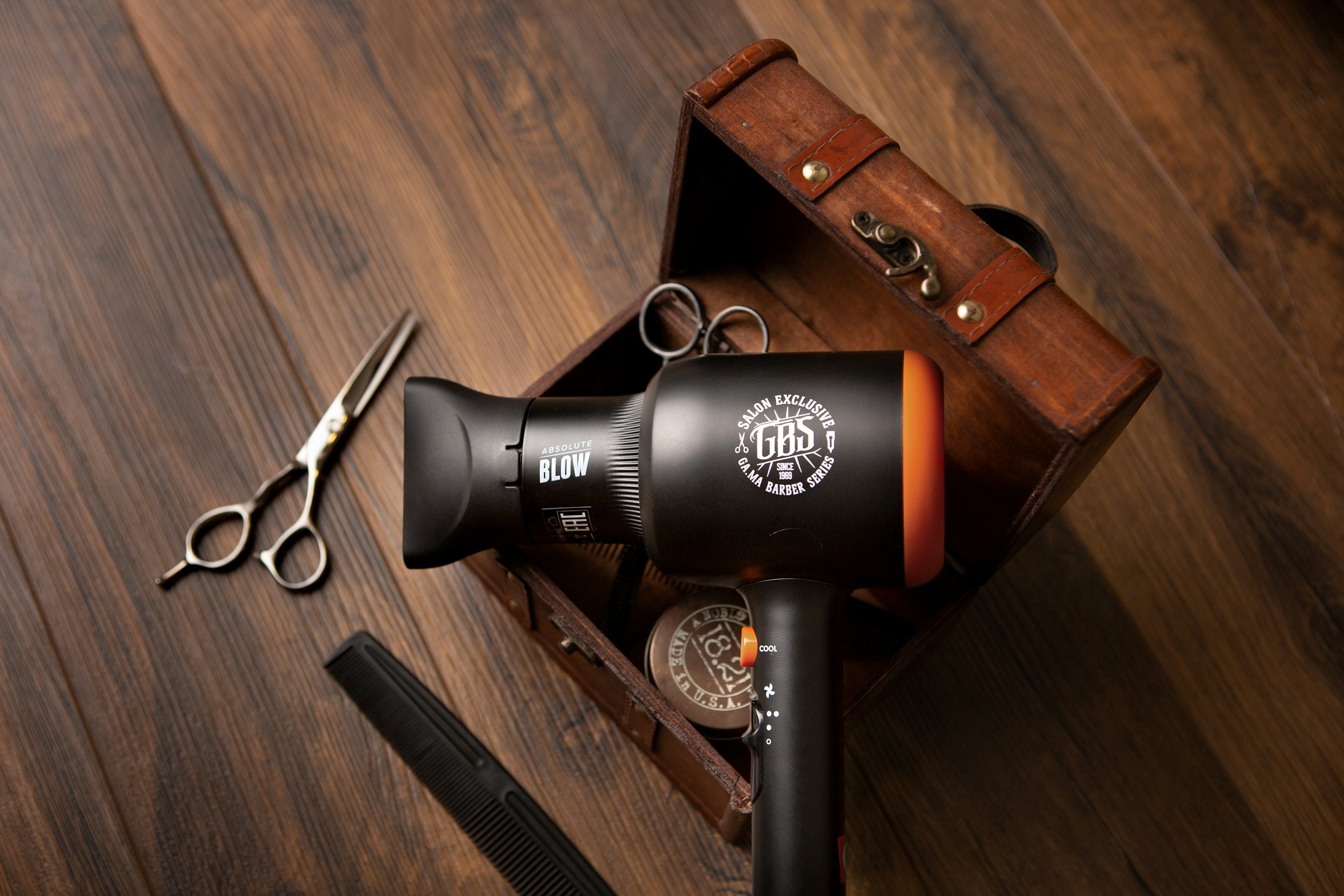 A wooden box containing a black and orange hair dryer, a pair of scissors, and a black comb, on a wooden floor.