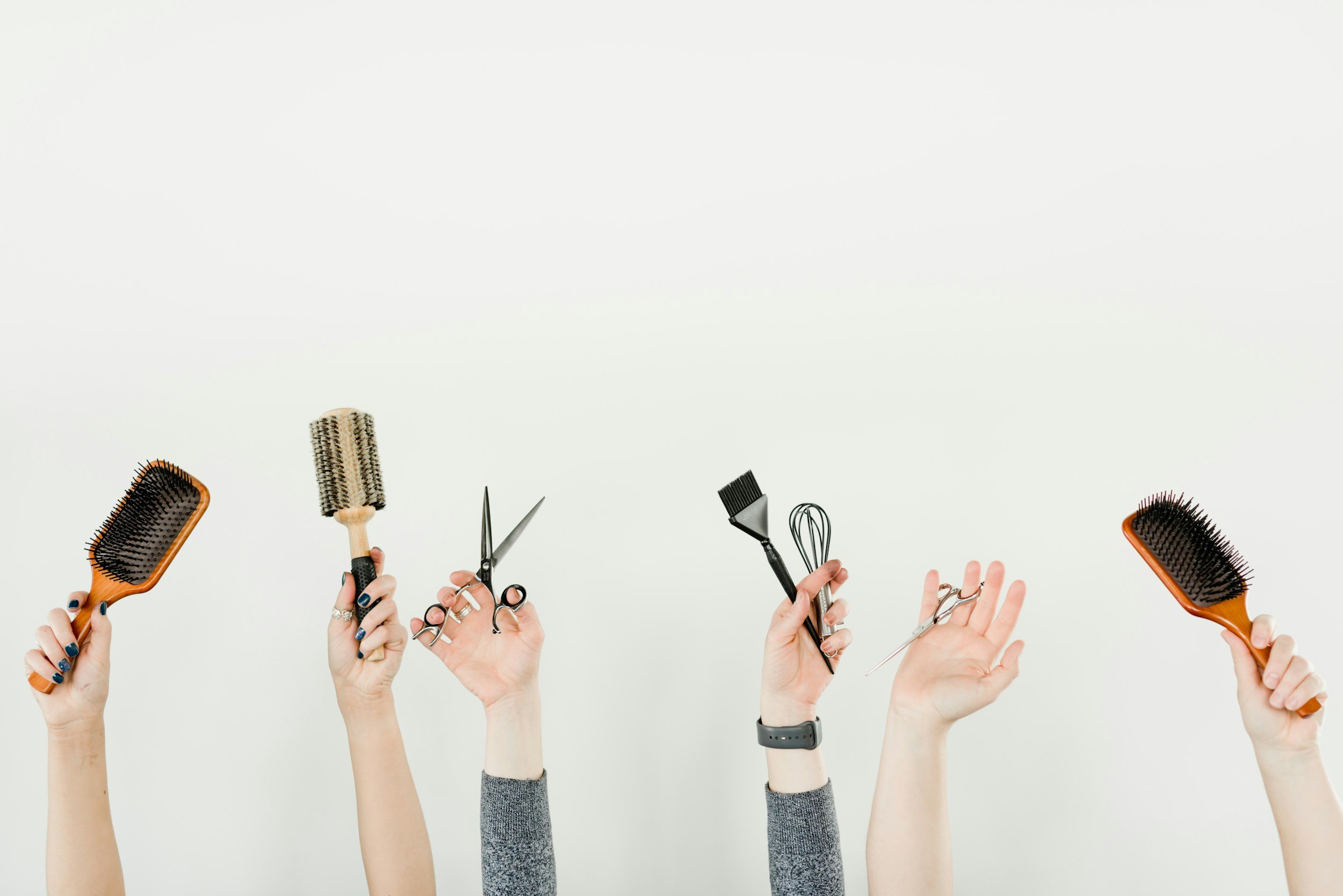 Multiple people's hands holding various hair styling tools including brushes, scissors, and a spray bottle against a plain white background.