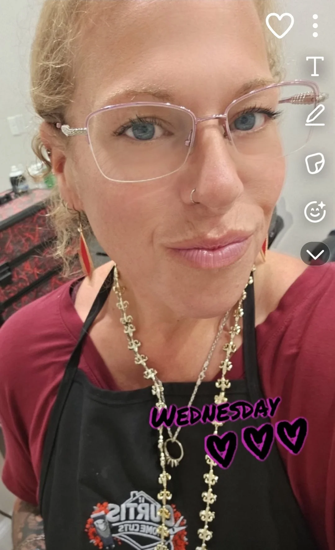 A woman with blonde hair, wearing glasses, a nose ring, and jewelry, taking a selfie in a room with a makeup or hair station in the background. She is wearing a maroon top and an apron with cartoon tools and the word "curtis" on it. The photo has text that says "Wednesday" with hearts.