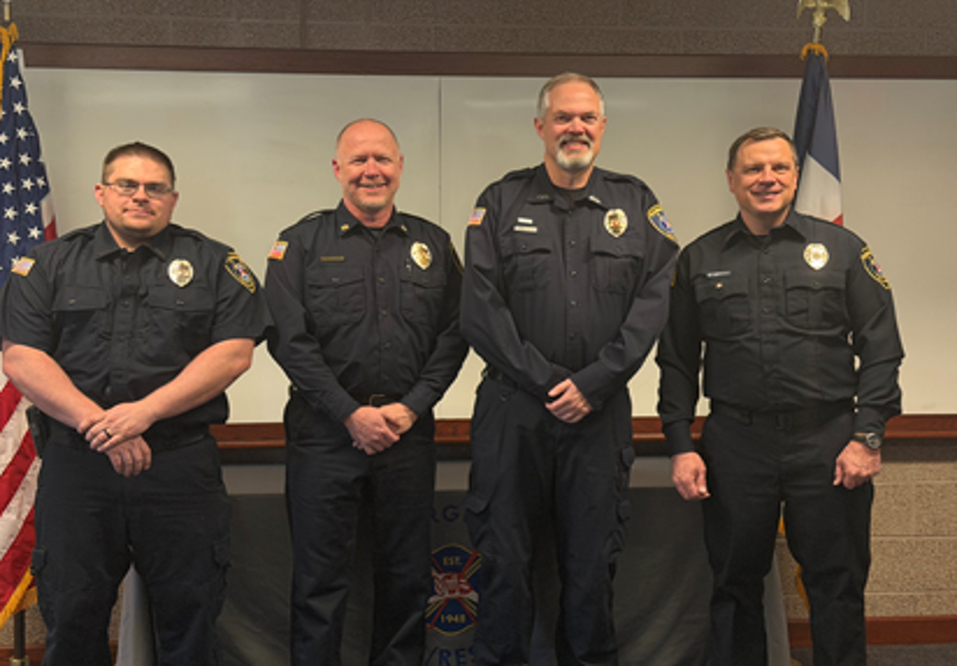 Four male police officers in uniform standing side by side in front of a whiteboard, with American and Texas flags behind them.