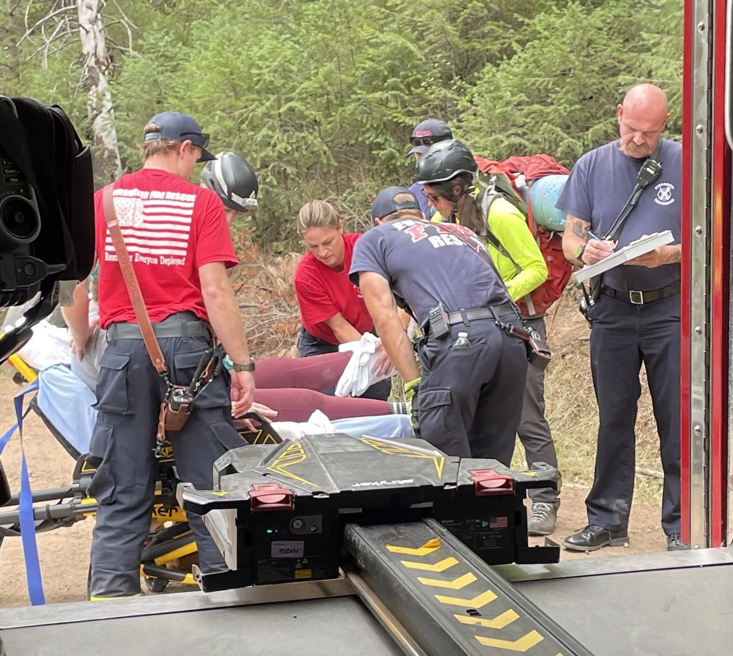 Emergency responders assisting a person on a stretcher in a forested area, with medical equipment nearby and an emergency vehicle visible.