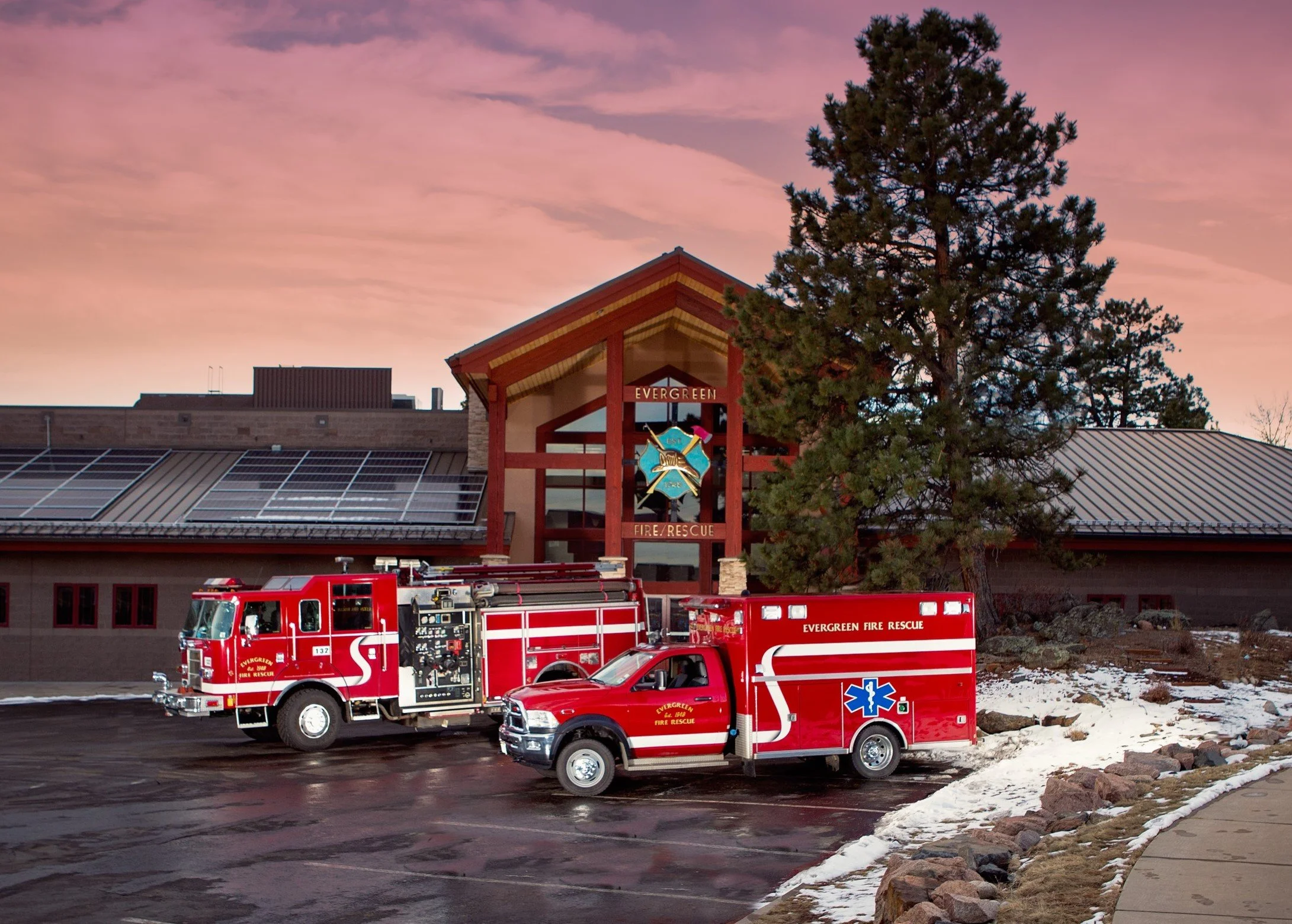 Emergency vehicles including fire trucks and an ambulance parked outside a building labeled 'Evergreen Fire/Rescue' at sunset with a large pine tree in the background.