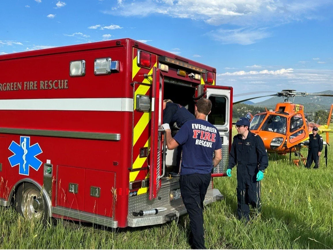 Fire rescue team members preparing rescue equipment from a red fire truck, with a helicopter in the background on a grassy field under a partly cloudy sky.