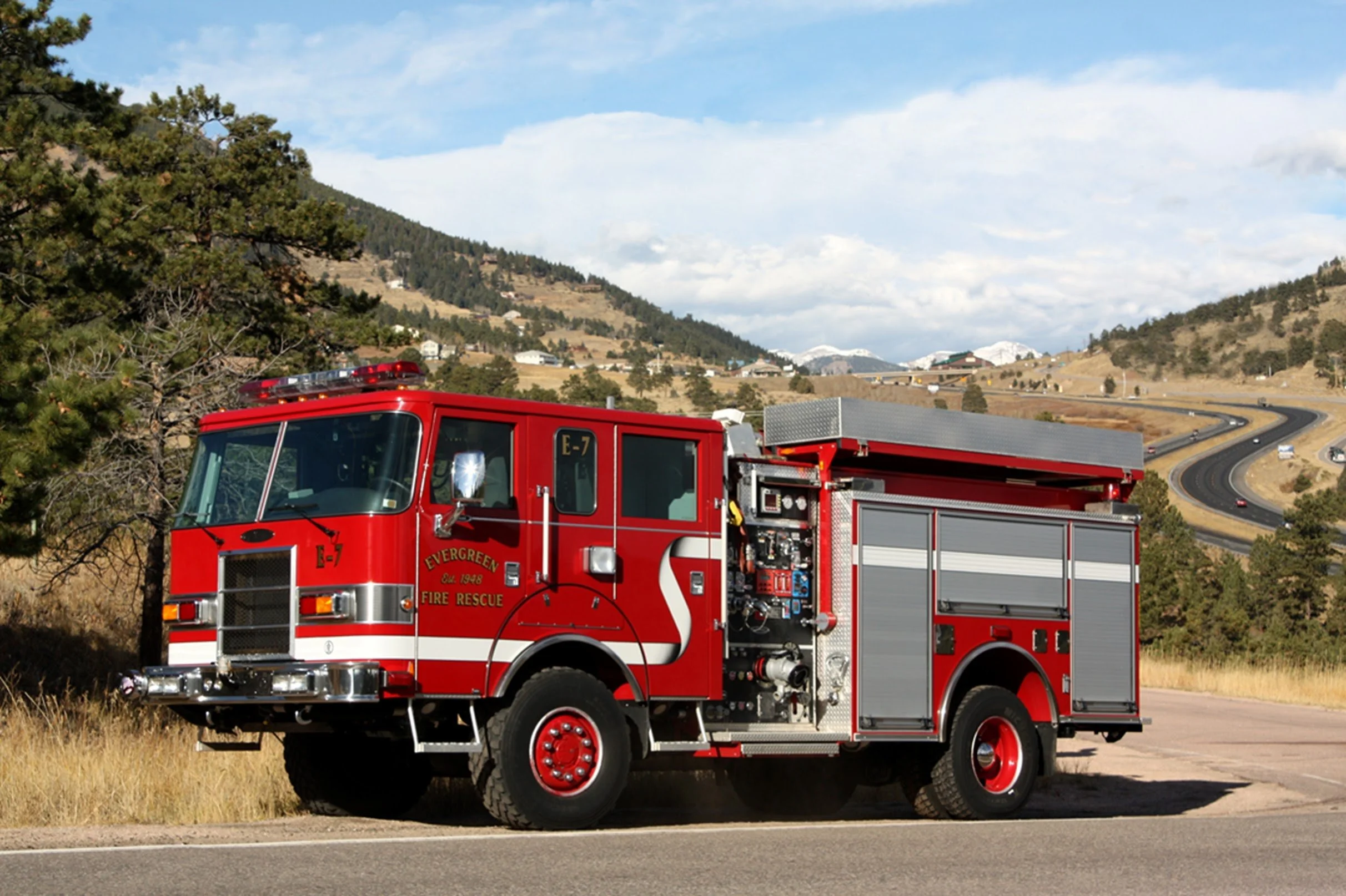 A red fire truck from Evergreen Fire Rescue parked on the side of a rural road with mountainous terrain and a partly cloudy sky in the background.