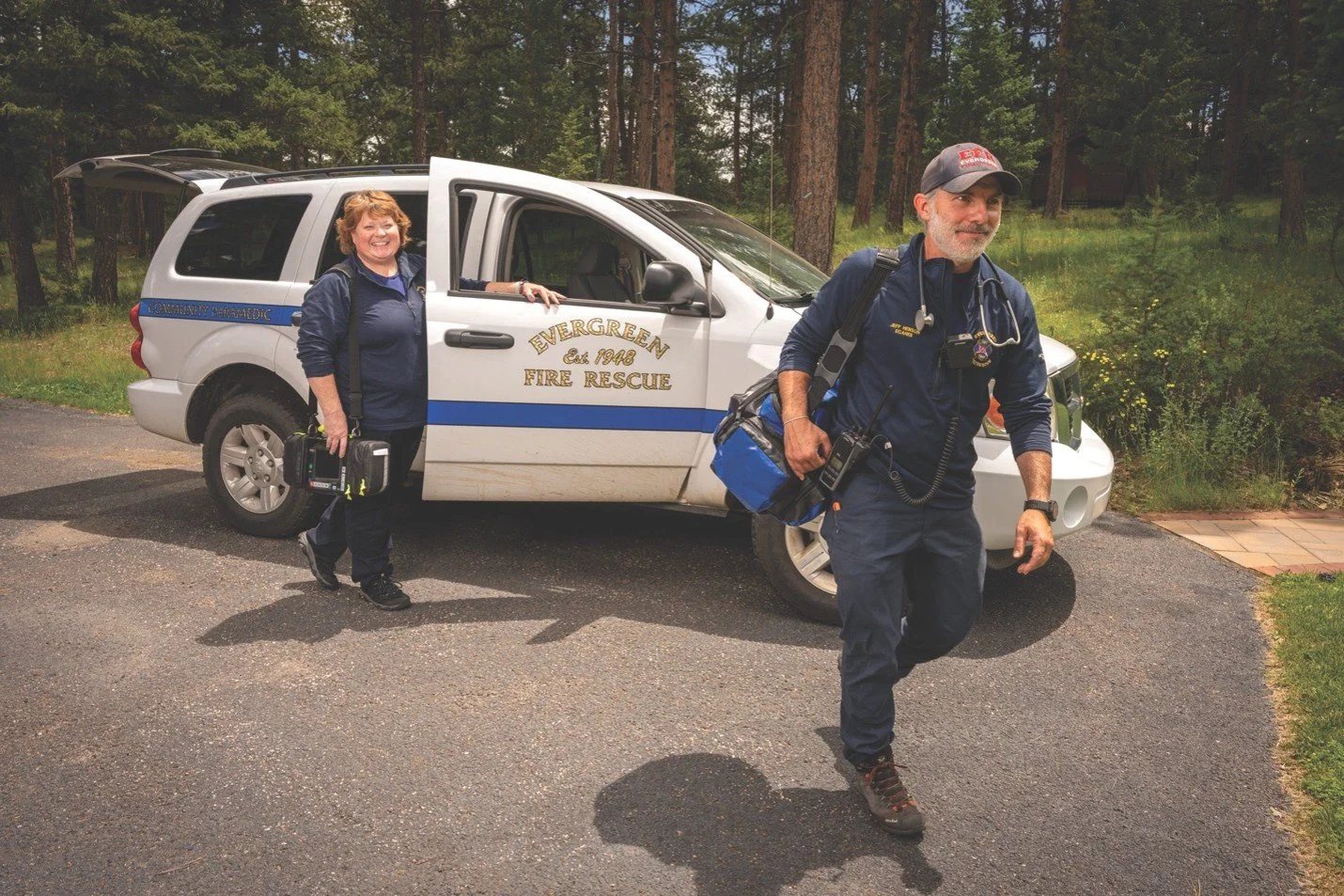 A male firefighter and a female emergency responder standing outside near an Evergreen Fire Rescue vehicle in a wooded area. The male firefighter has a beard, wears a cap, and has emergency gear. The female emergency responder is smiling, holding equipment, and standing partially inside the vehicle.