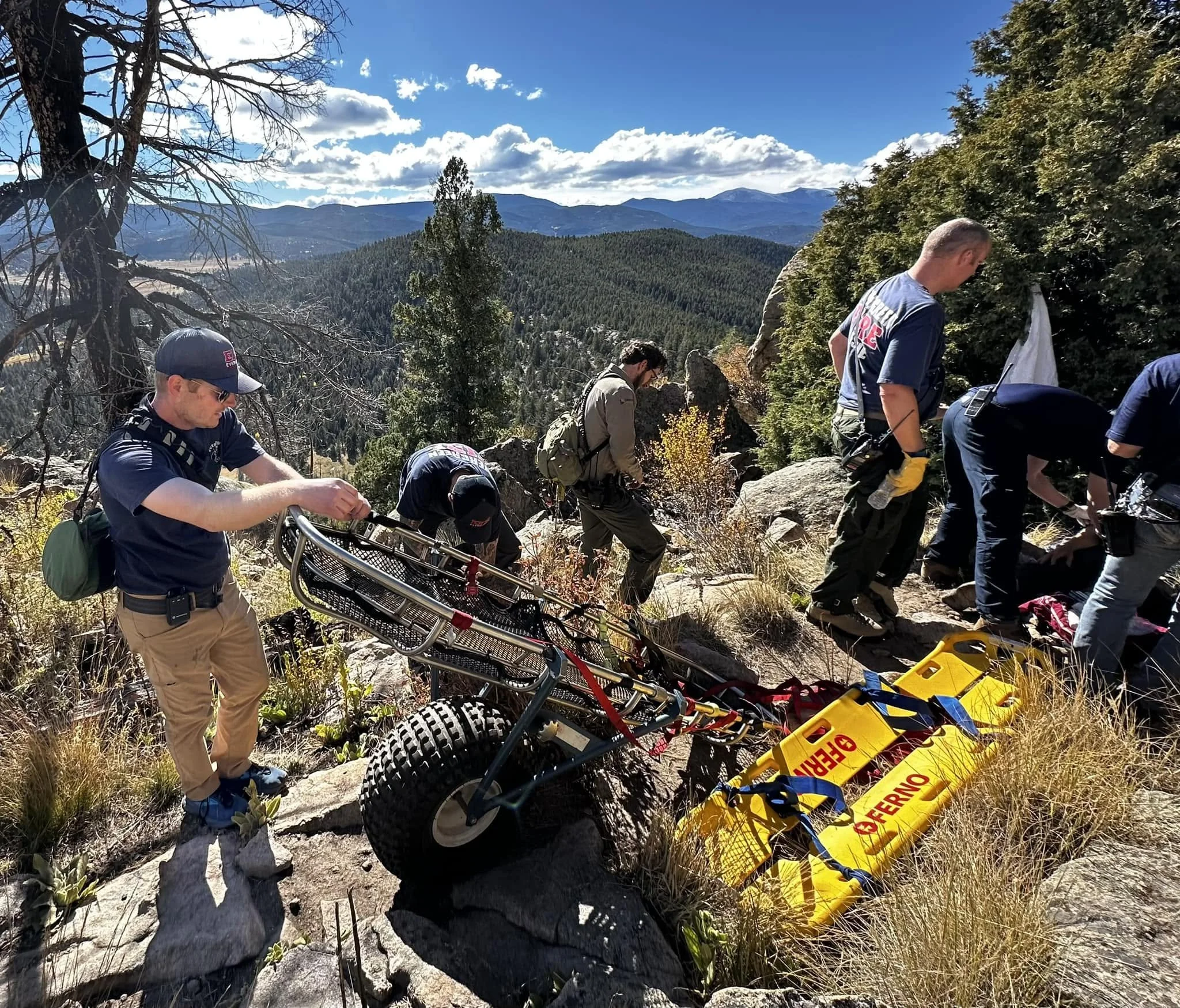 Group of people on a mountain trail with emergency medical equipment, including a stretcher and caution tape, in a scenic landscape with mountains and blue sky.