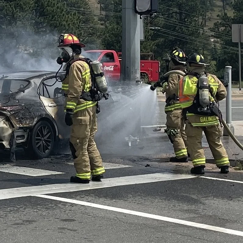 Three firefighters in full gear extinguishing a car fire at an intersection. The car has visible damage and smoke coming from it.