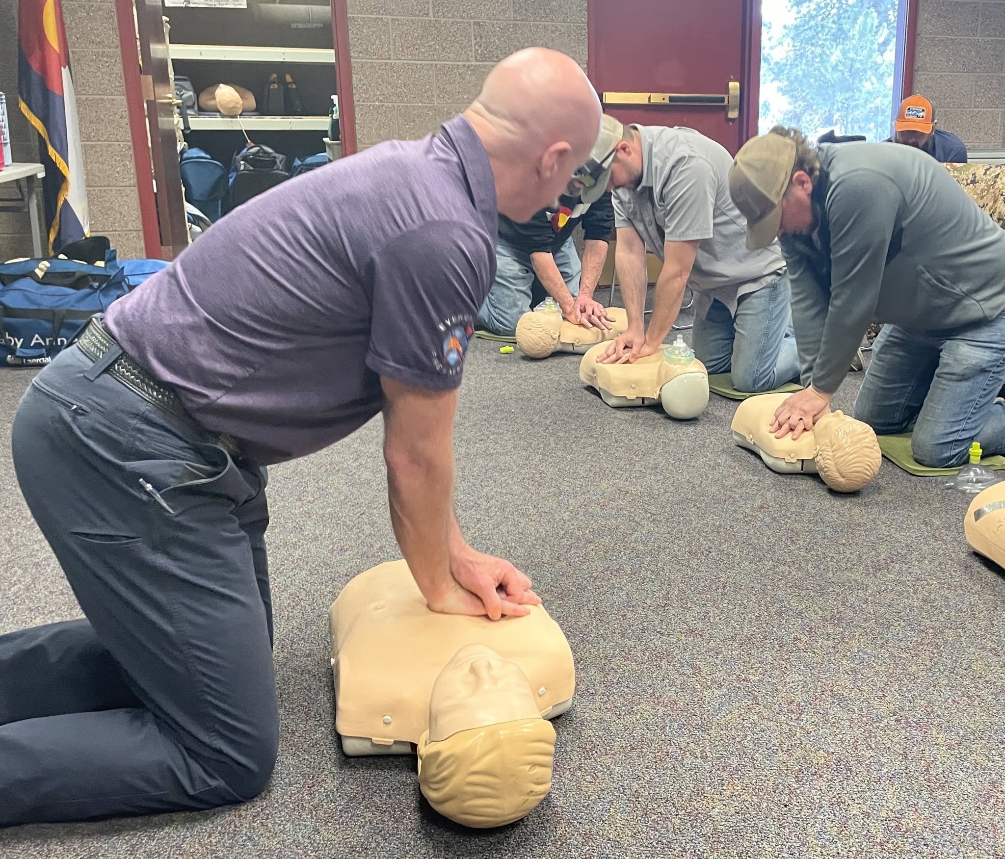 A group of men practicing CPR on mannequins during a training session in a room with backpacks on shelves and a flag on the wall.
