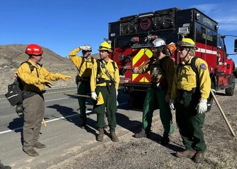 Six emergency responders, four men and two women, wearing yellow and green uniforms with helmets, standing next to a red and black fire truck on a roadside in a desert area, during daytime.