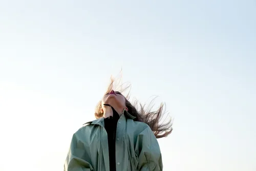 A woman with long hair blowing in the wind, wearing a jacket, standing outdoors against a clear sky.