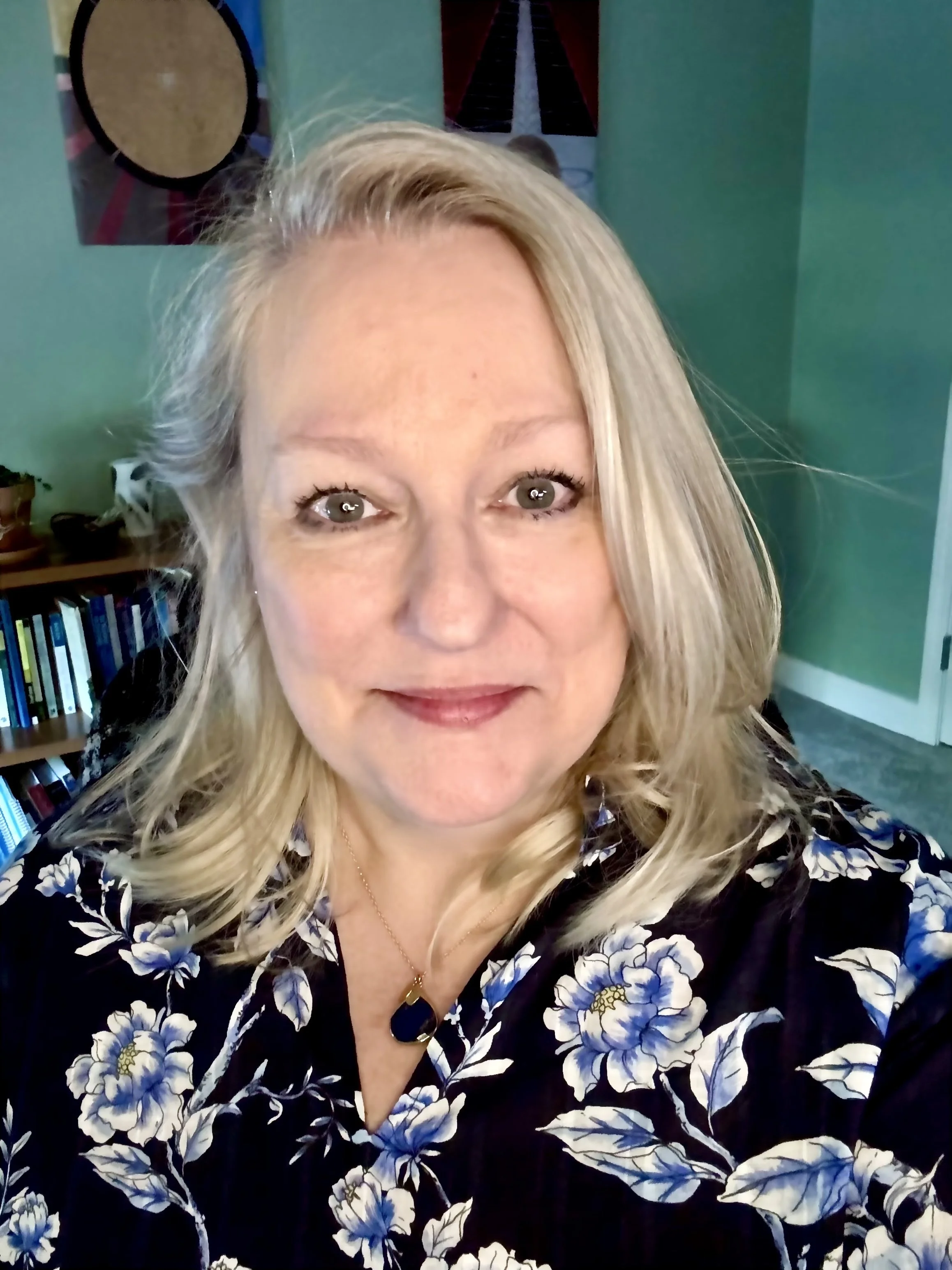 Close-up of a woman with long blonde hair, light skin, and gray eyes, wearing a black floral blouse with blue and white flowers and a gold necklace, indoors with a teal wall and artwork in the background.