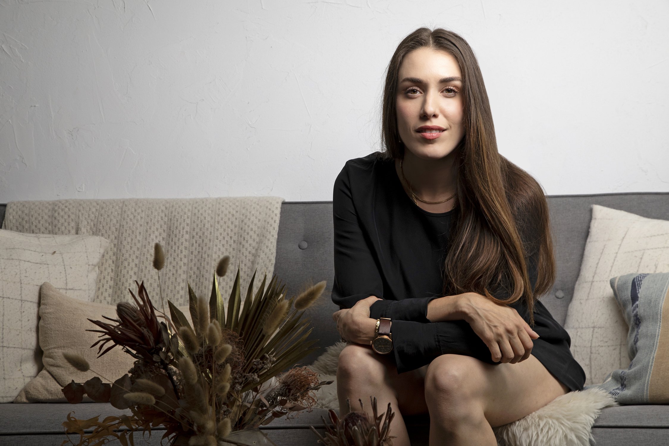 A young woman with long brown hair, wearing a black top and jewelry, sitting on a couch with beige and neutral colored pillows, in front of a white textured wall, with plants and decor in the foreground.