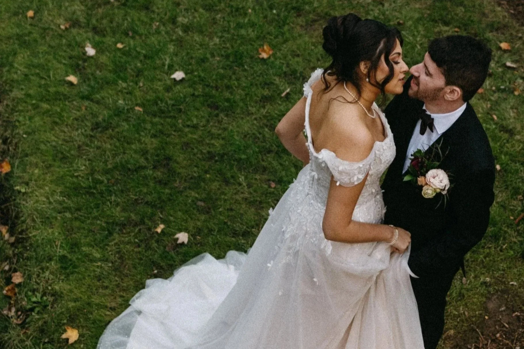 A bride and groom sharing a close moment outdoors on a grassy area with fallen leaves, the bride in a white off-shoulder wedding dress and pearl jewelry, and the groom in a black tuxedo with a boutonniere.