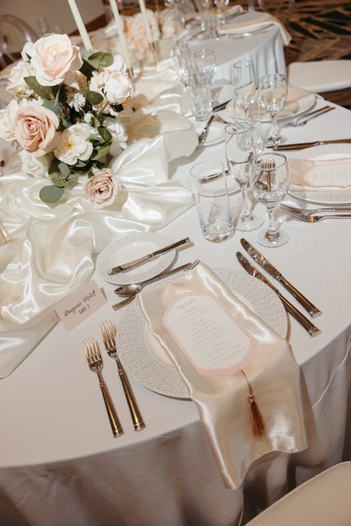 Elegant banquet table decorated with a white satin tablecloth, a floral centerpiece with roses, and place settings including silverware, glassware, and a printed menu on a silk napkin.