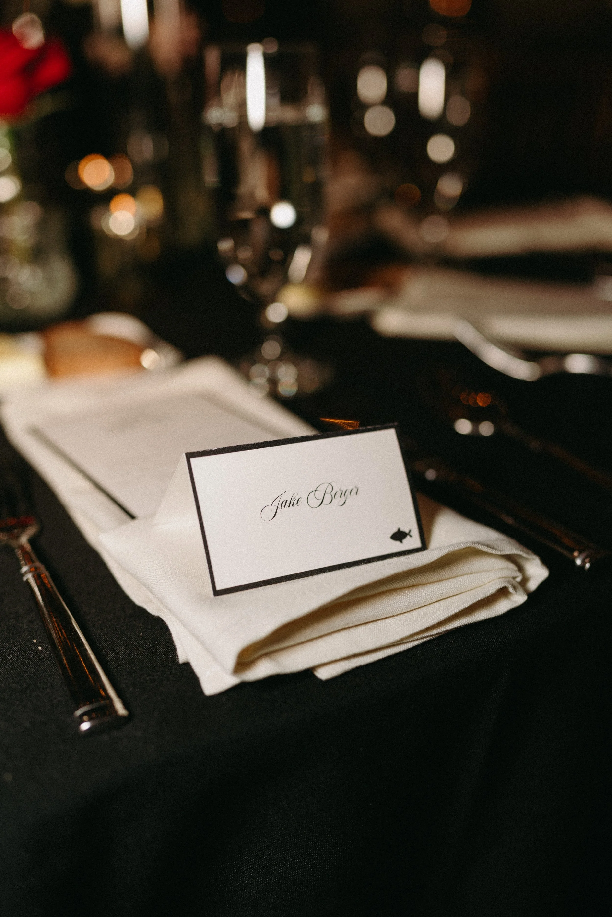 A table with a name card reading 'Jake Berg'. The table is set with a folding napkin, a pen, and other dining utensils. In the blurred background, there are glasses and a person's hand.