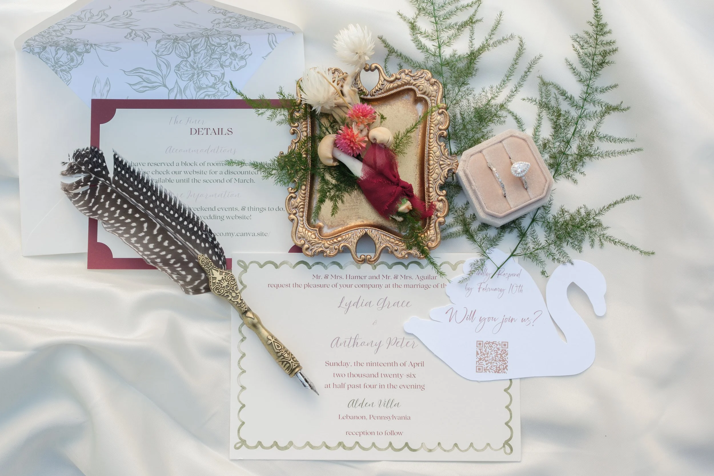 A wedding invitation surrounded by greenery, a decorative frame with artificial flowers, a feather, and a ring in a jewelry box.