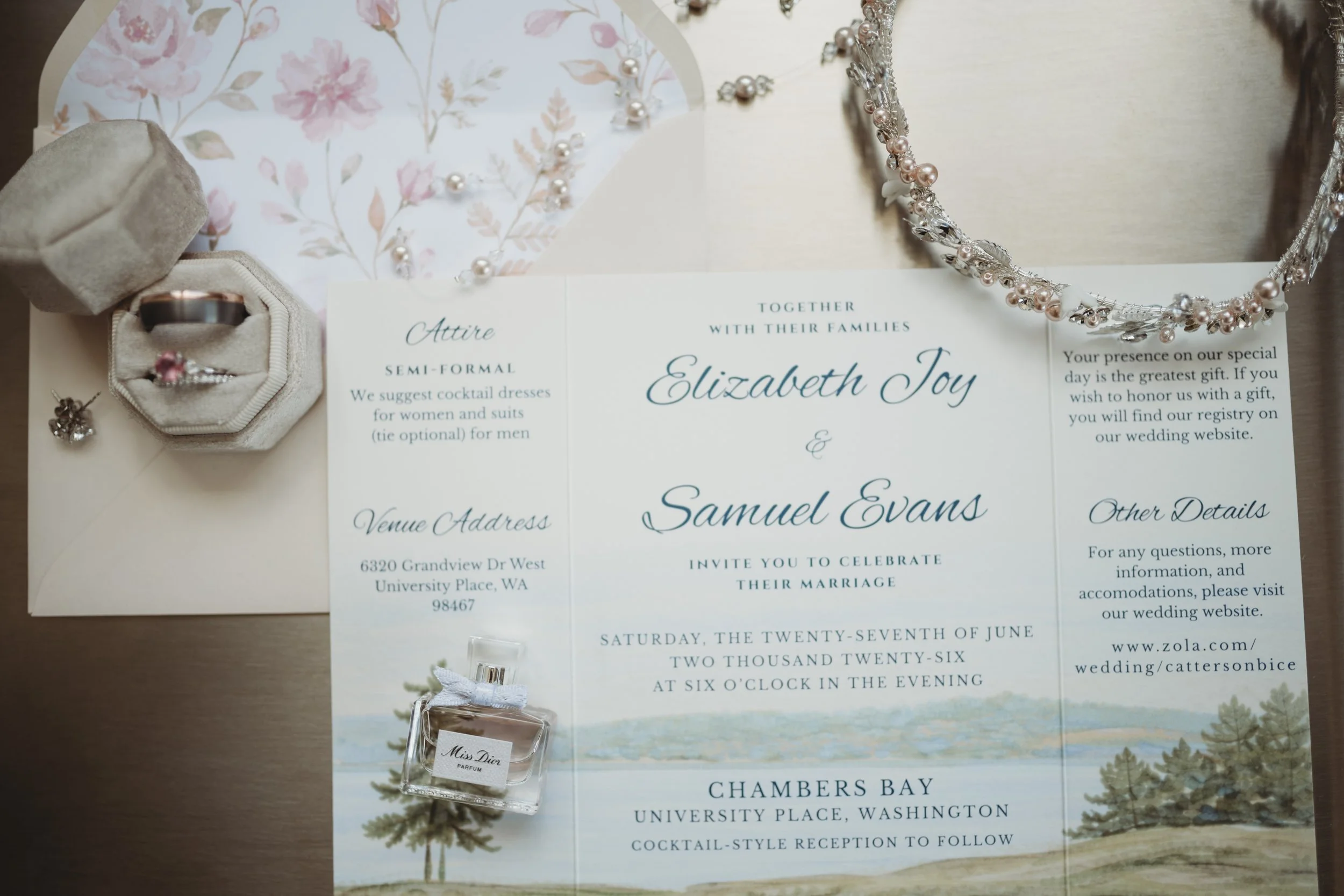 Wedding invitation, jewelry including a necklace and earrings, a ring in a box, and a perfume bottle on a table.