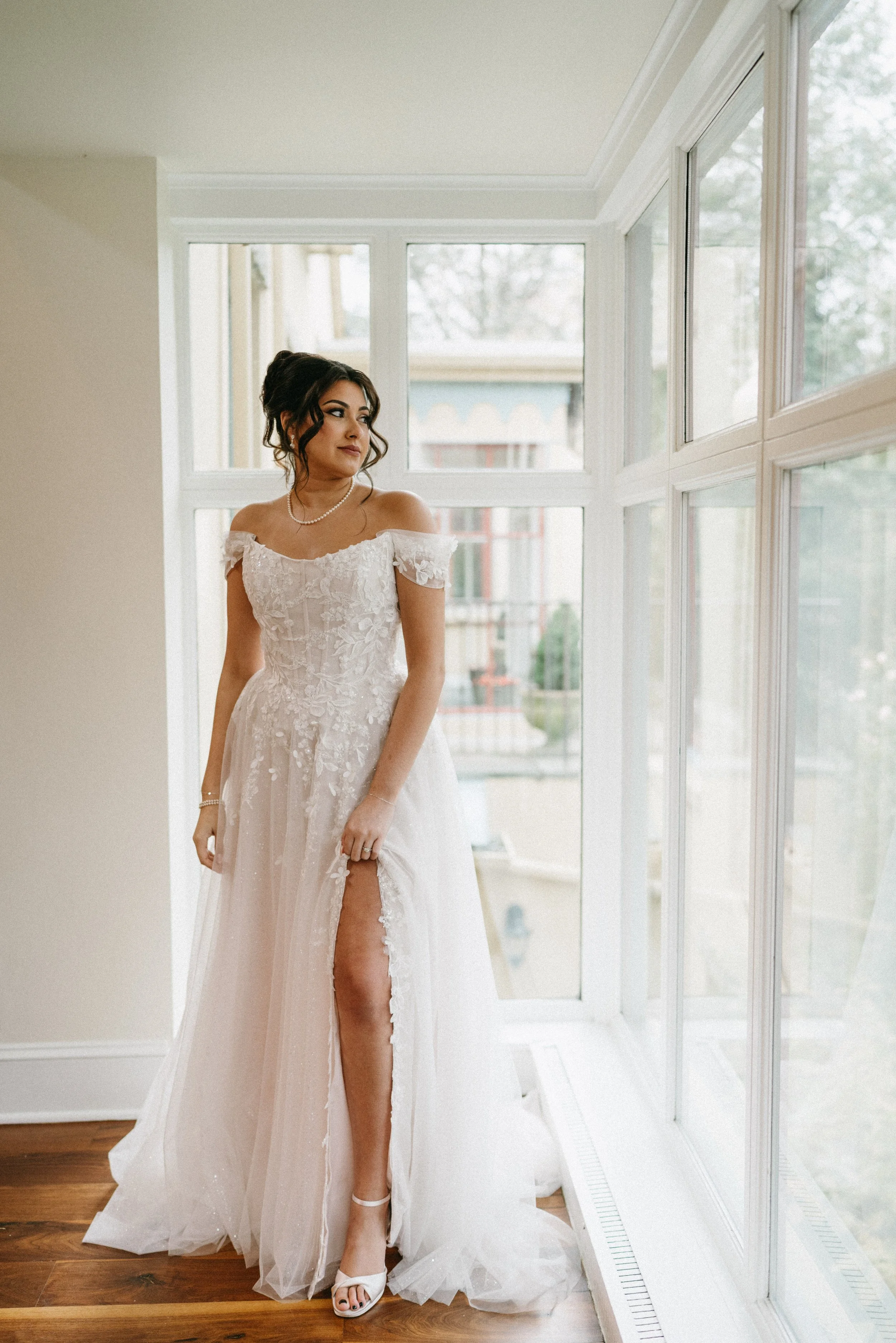A bride standing indoors near large windows, wearing a white lace off-shoulder wedding gown with a thigh-high slit, white high heels, a pearl necklace, and earrings, with her dark hair styled in loose curls.