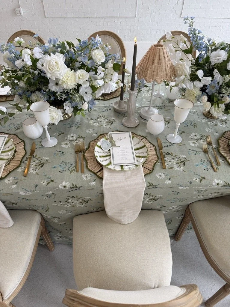 A beautifully decorated dining table set for a formal event with floral centerpieces, candles, and elegant tableware, featuring a floral tablecloth and a small menu card.