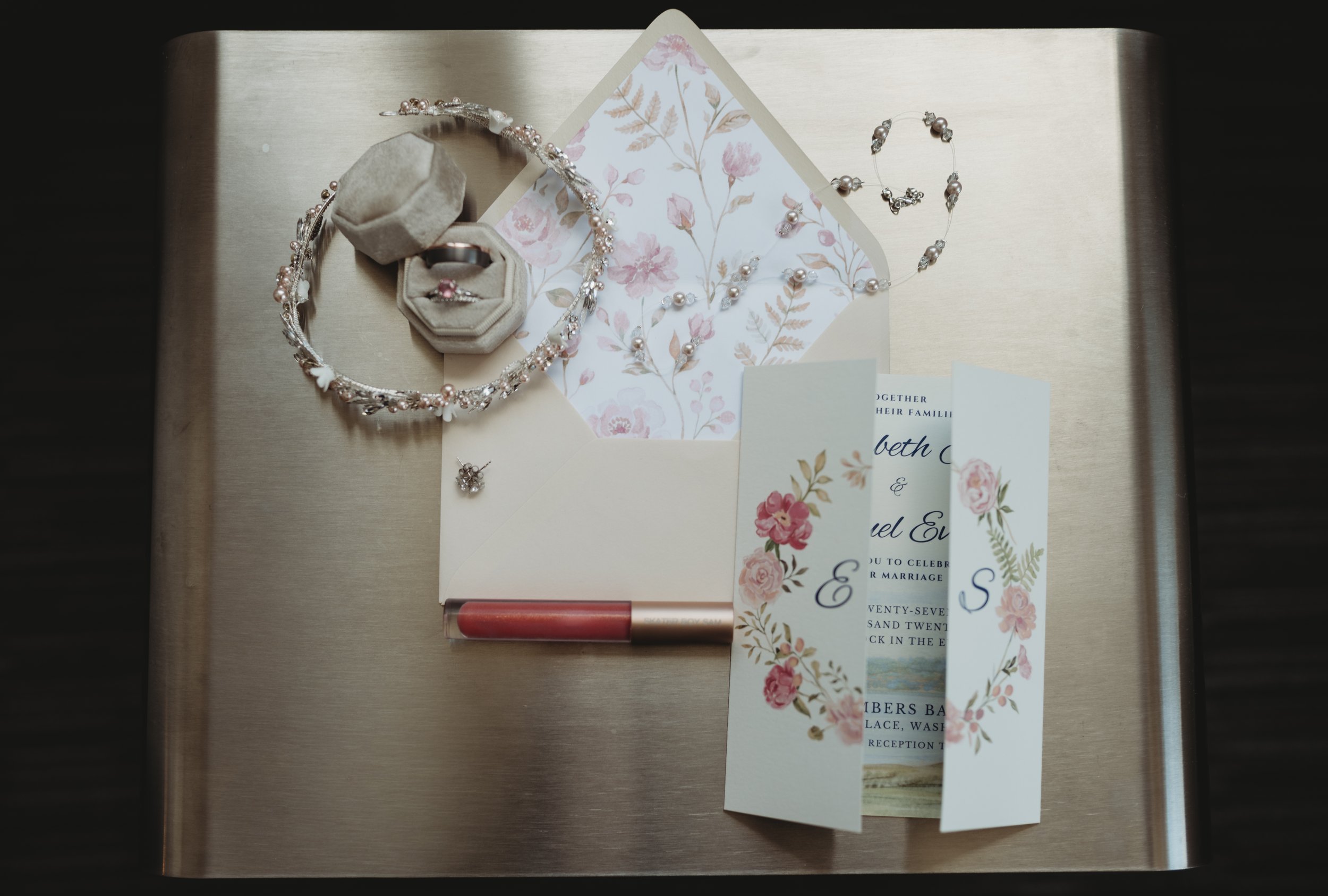 A collection of wedding jewelry and items on a metallic surface, including a ring in a beige ring box, a pearl necklace, a pair of earrings, and wedding invitations with floral designs.