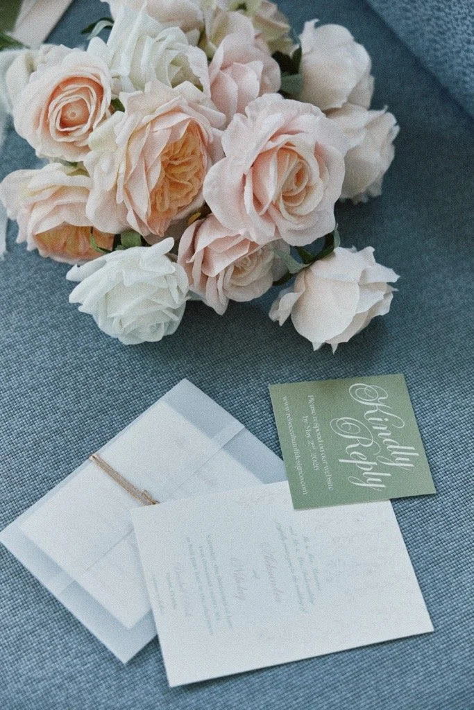 A bouquet of light pink and white roses on a gray surface next to wedding invitations and a green card reading "Wedding & Happy".
