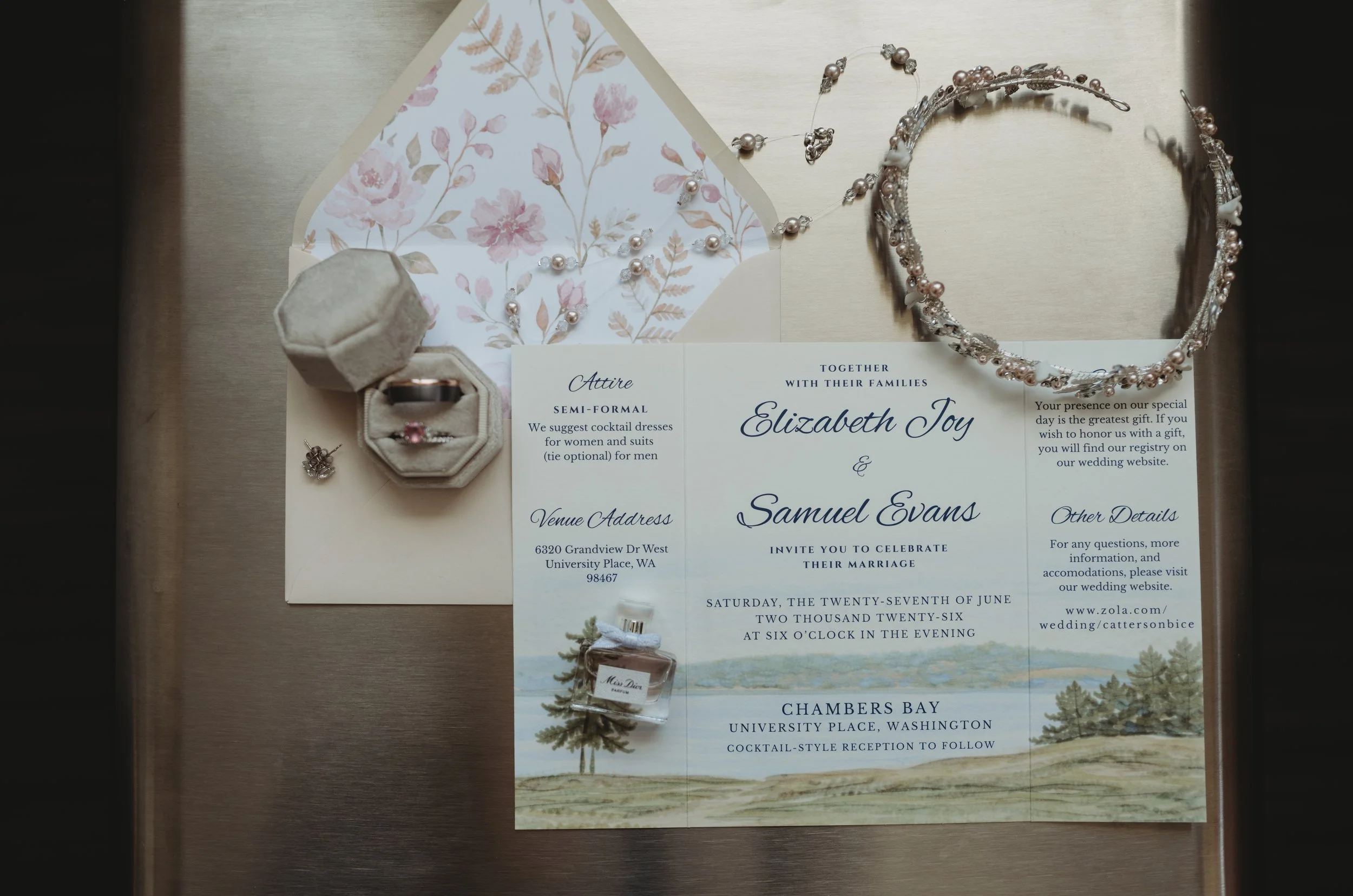 Wedding invitation card with floral design, jewelry including a pearl necklace and earrings, a ring in a velvet box, and a small perfume bottle, all arranged on a wooden surface.
