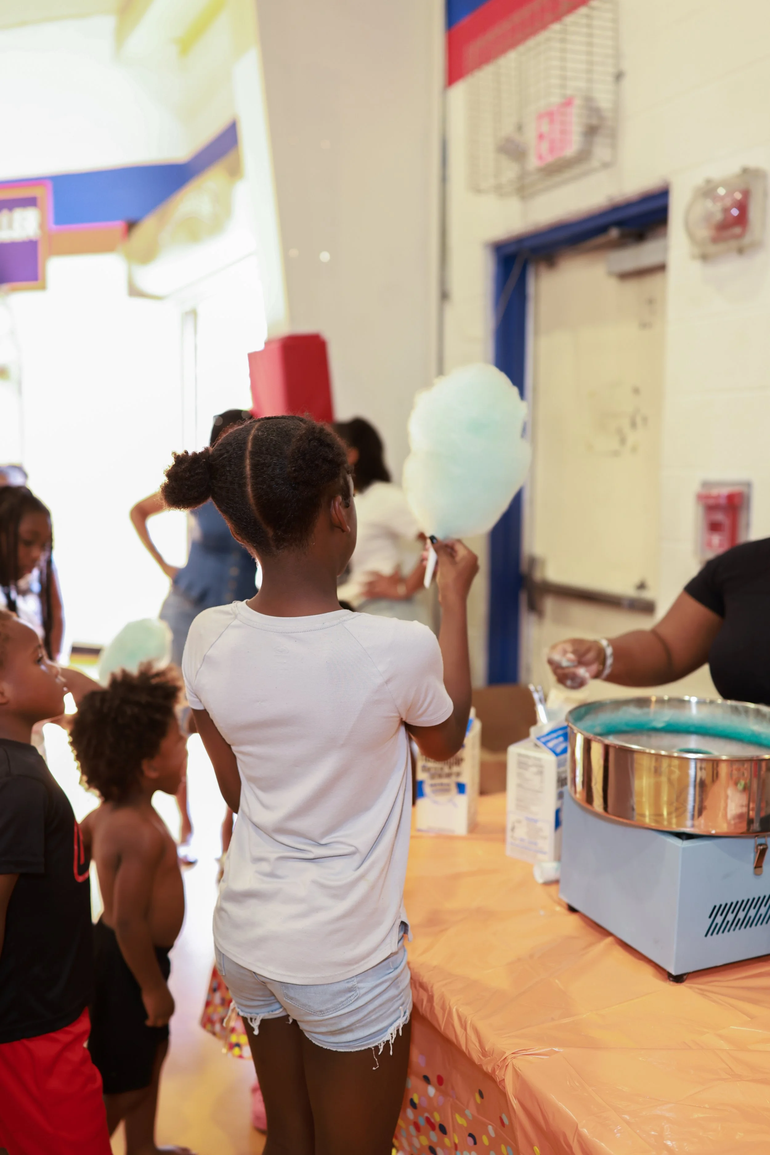 Children standing in line at a carnival or fair, waiting for cotton candy from a vendor. The vendor's table is covered with a colorful, polka-dotted tablecloth, and a cotton candy machine is visible in the foreground.