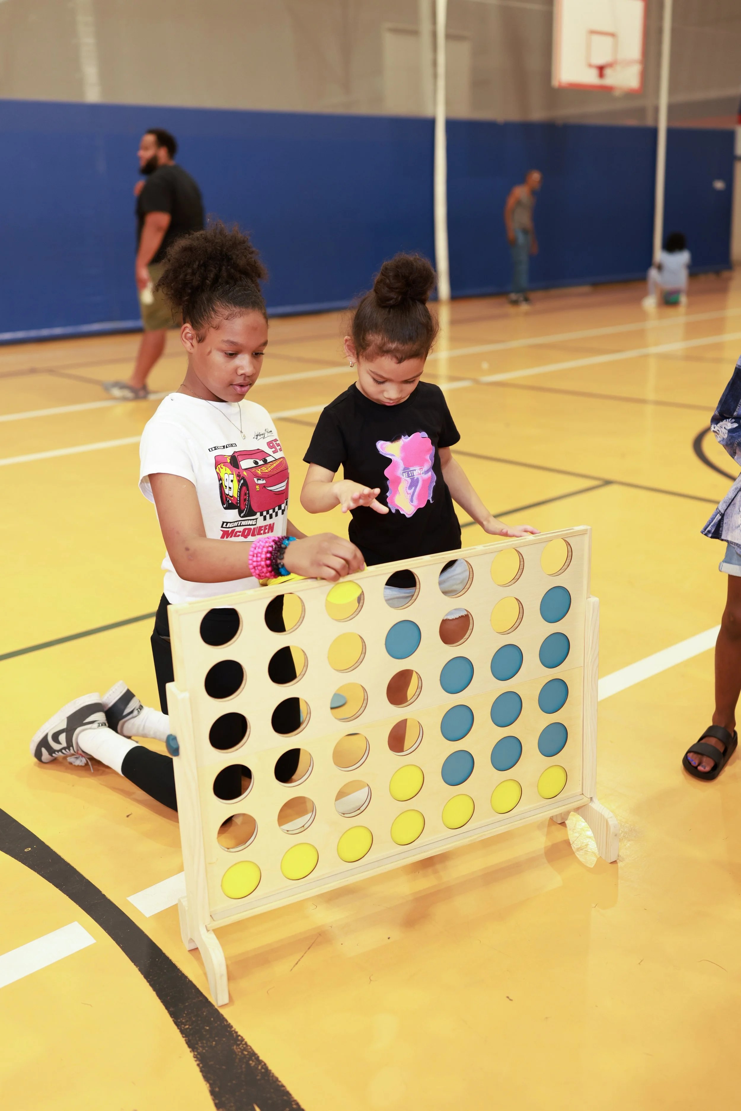Two young girls are kneeling on the floor of a gym, playing Connect Four. The girl on the left is wearing a white Lightning McQueen T-shirt and has her hair in a ponytail, while the girl on the right is wearing a black T-shirt with a unicorn design a