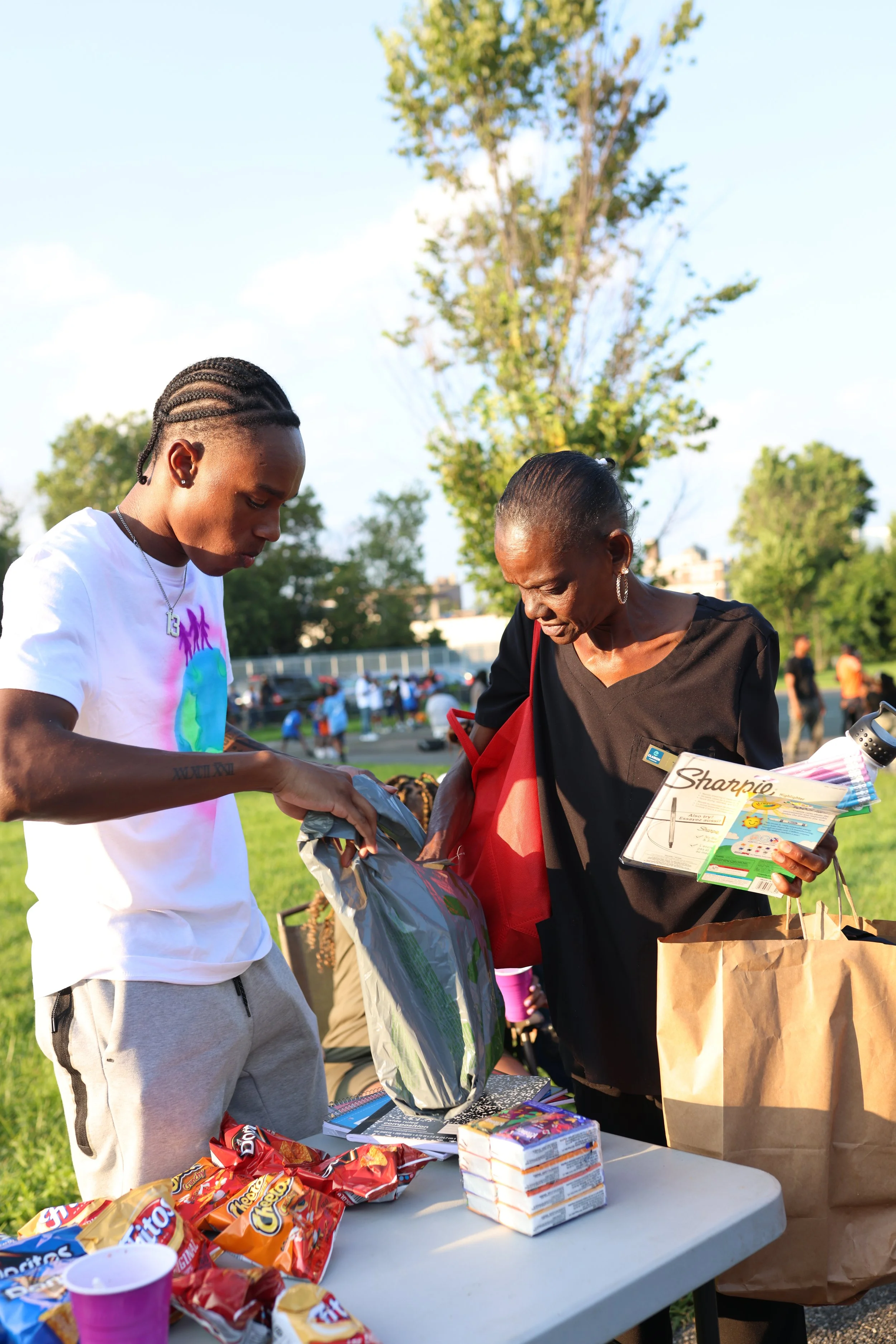 Two women are at a table outdoors with snacks and boxes of food, engaging in conversation during what appears to be a community gathering or event.