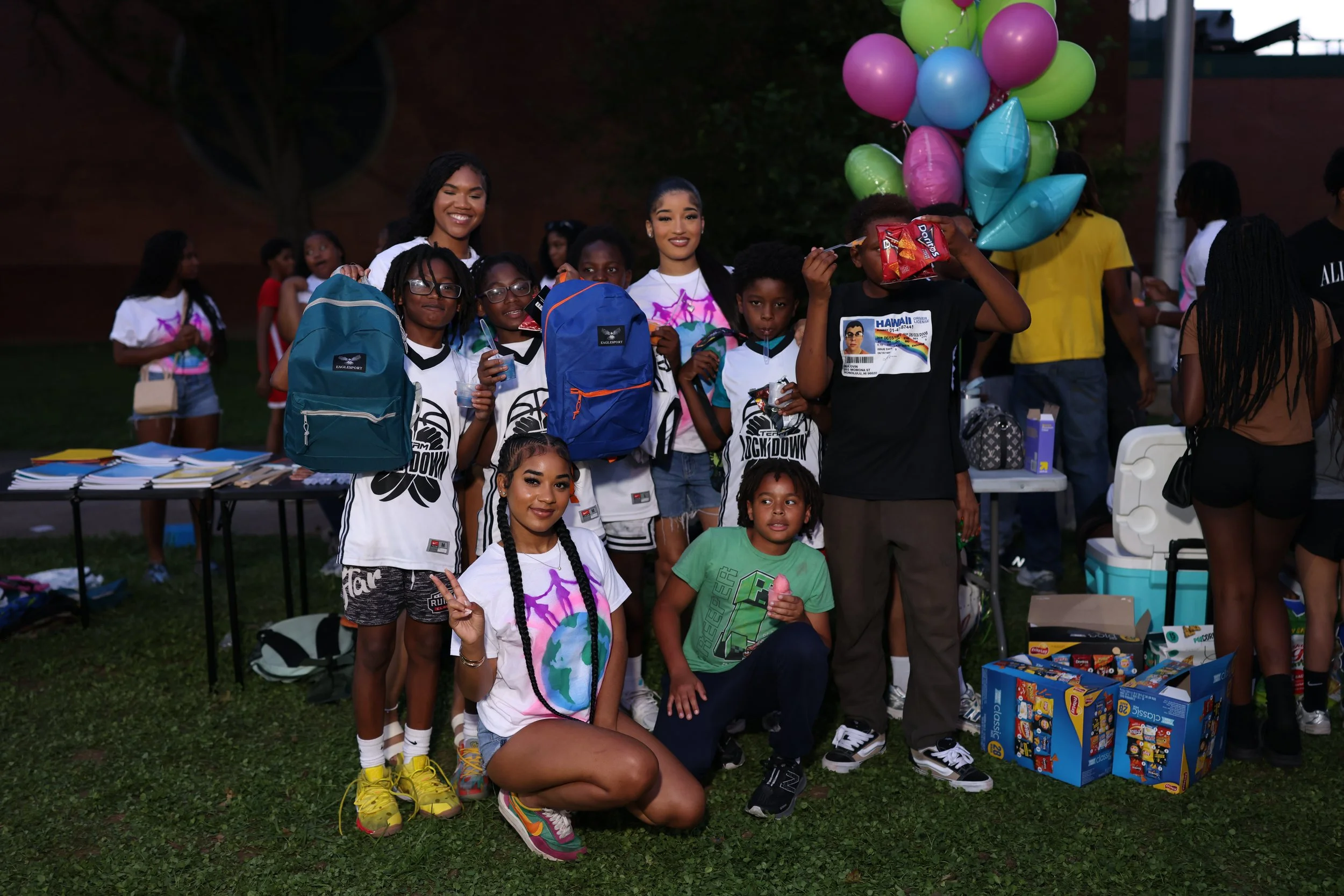 A group of children and young adults gathered outdoors at night, some holding balloons and snacks, with tables and boxes of toys in the scene.