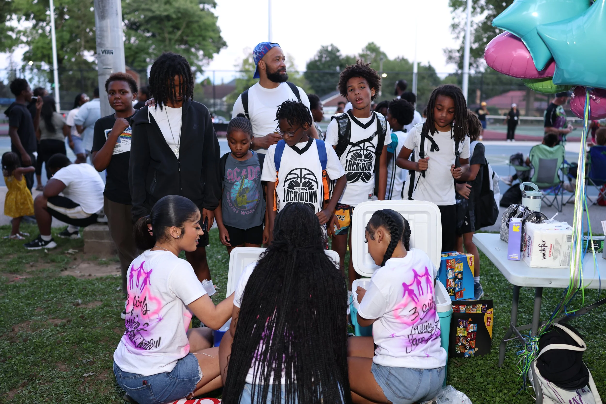 A group of children with backpacks and some wearing matching tie-dye T-shirts are gathered around a table at an outdoor event. In the background, there are more people, balloons, and a sports court with trees and a few houses visible.