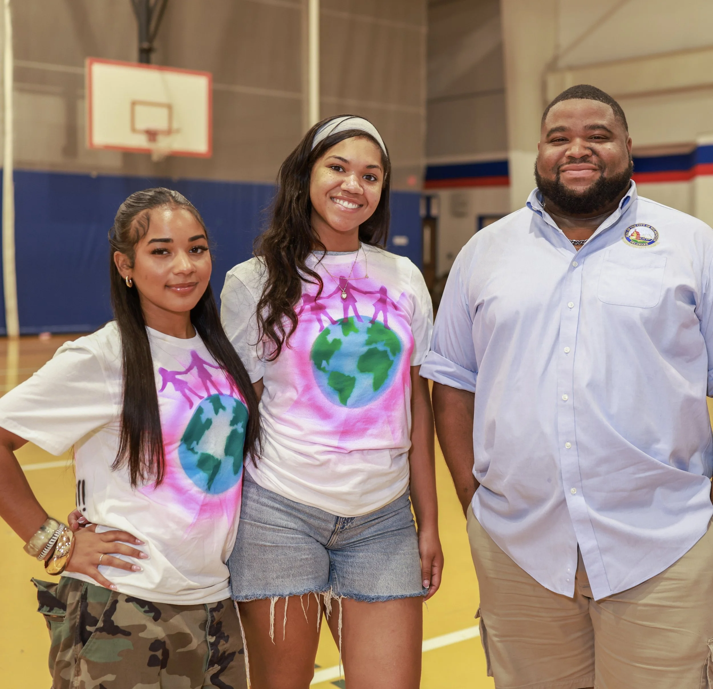 Three people standing in a gymnasium, smiling and posing for the photo. The person on the right is wearing a white shirt with a badge, the woman in the middle is wearing a tie-dye shirt with a world map design, and the girl on the left is wearing a w