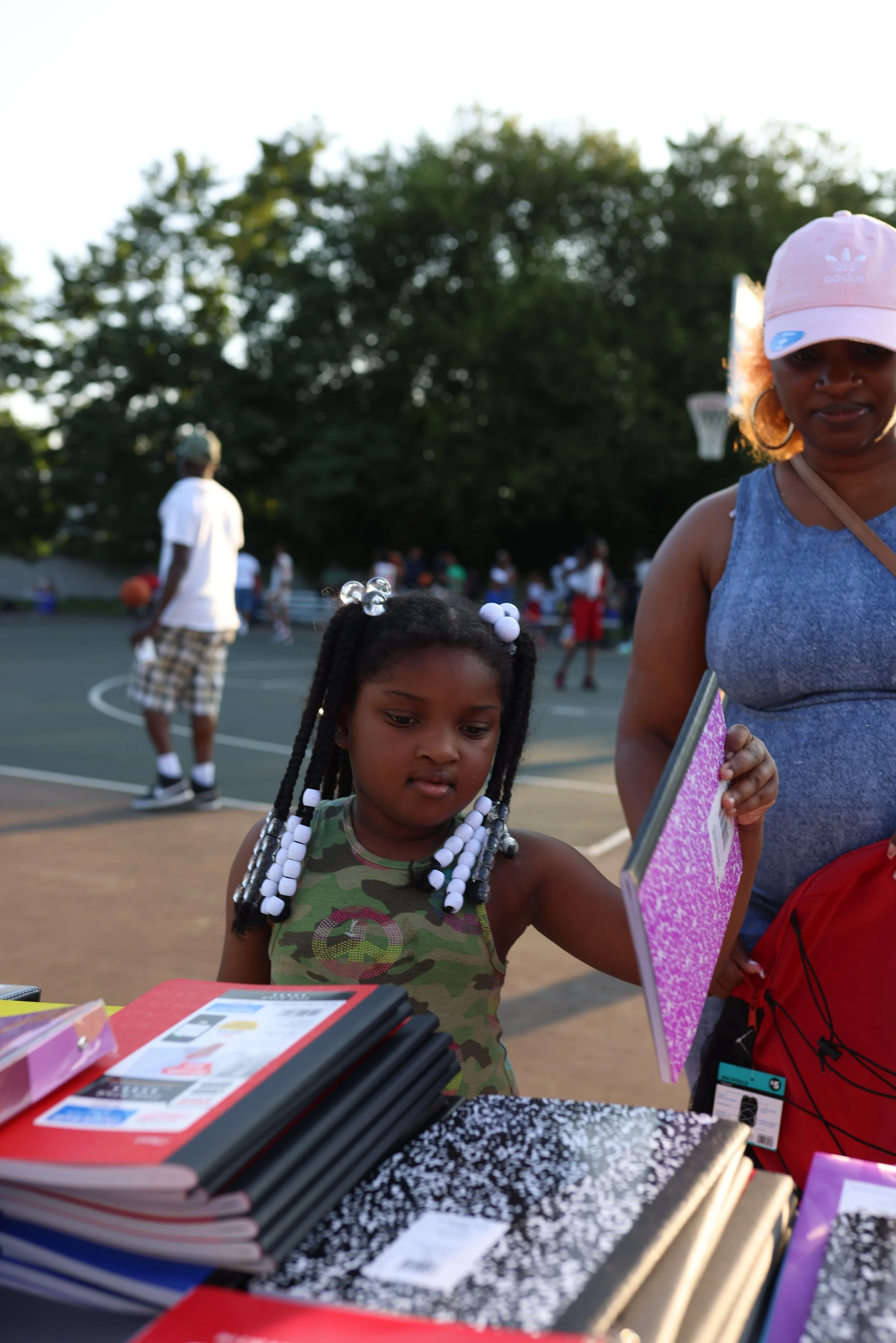 A young girl with braided hair and beads wearing a camouflage shirt, and a woman wearing a pink Adidas cap, blue tank top, and hoop earrings, standing outdoors at a table with notebooks and school supplies, during daytime with a basketball court and 