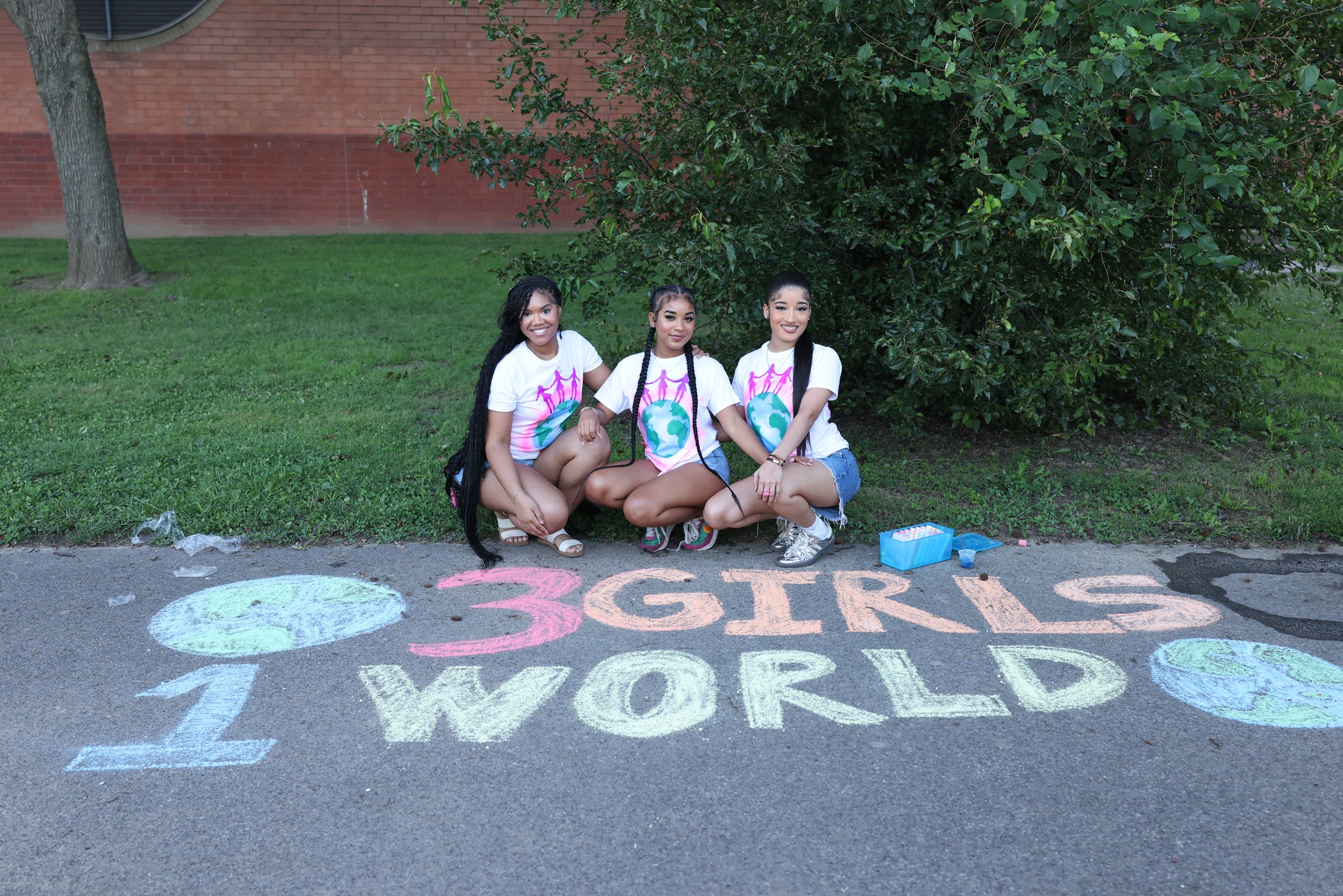 Three young women sitting on the sidewalk under a tree, wearing matching white t-shirts with a globe and pink figures, and colorful shorts. They are smiling at the camera, and there are chalk drawings and writing on the ground that says "3 GIRLS" and