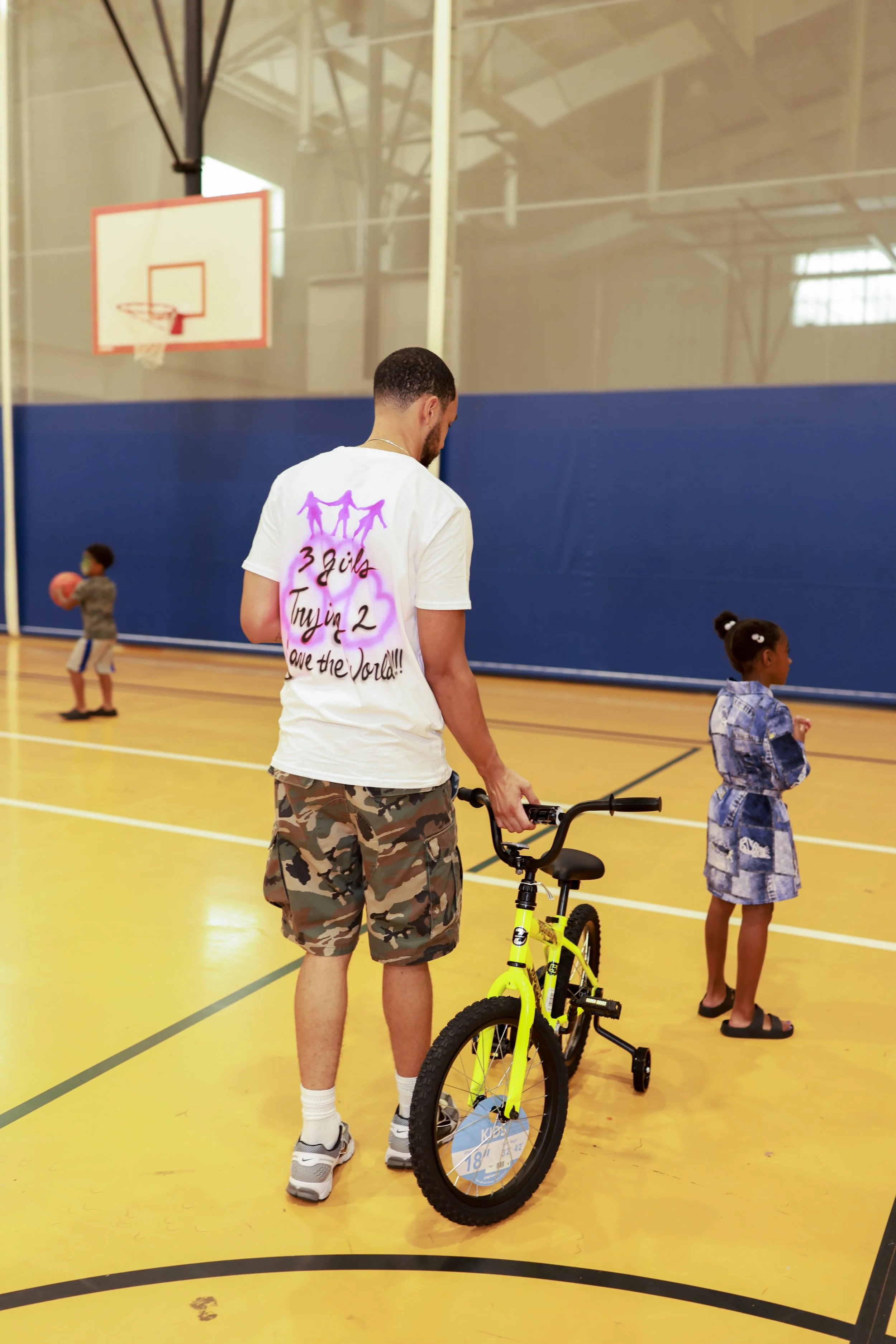 A man with a beard wearing a white t-shirt and camouflage shorts stands in a gymnasium, holding a yellow bike. There are children playing in the background near a basketball hoop.