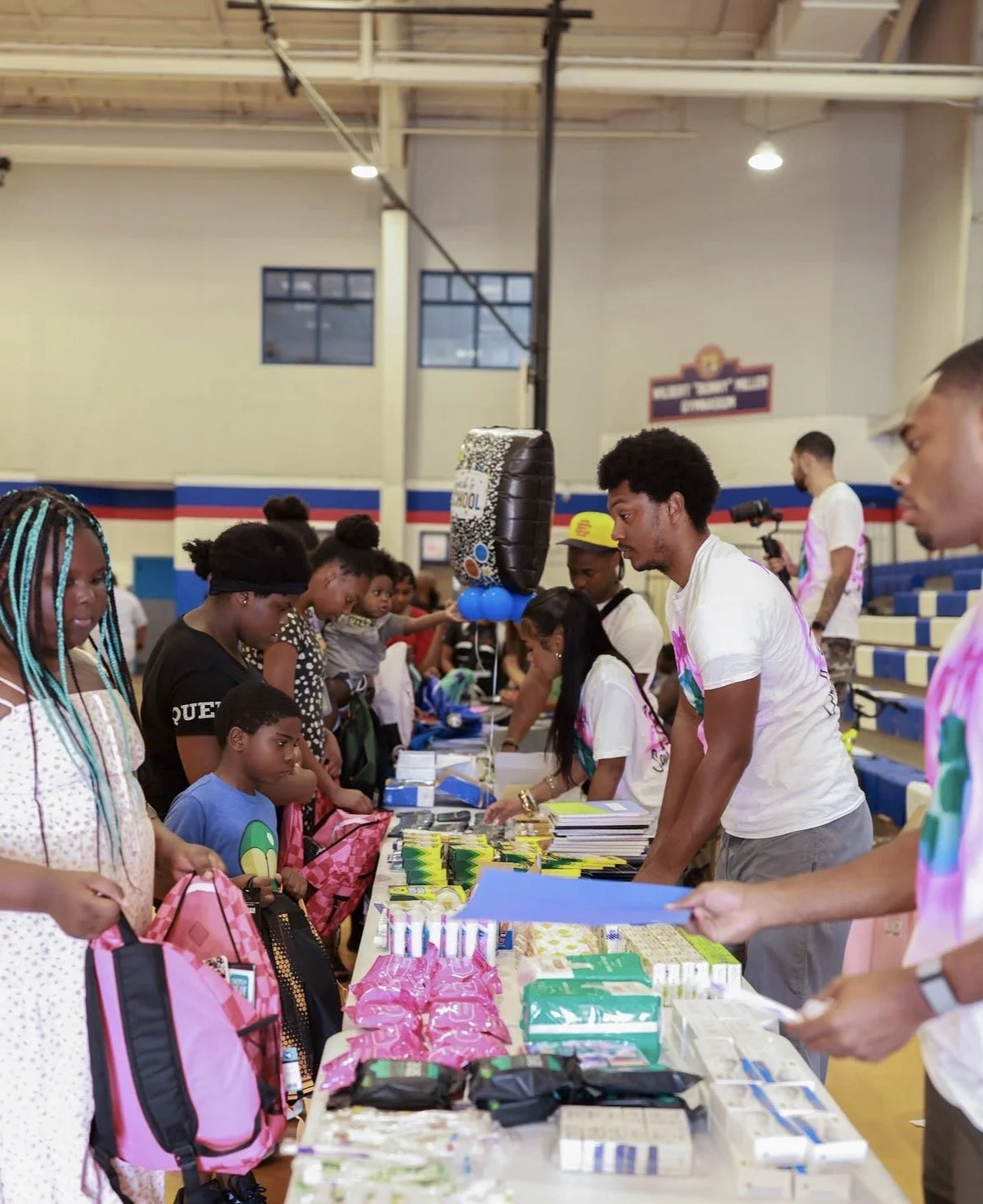 People interacting at a table filled with various items, likely at a community event or donation drive inside a gymnasium with blue and yellow seating and a balloon decoration.