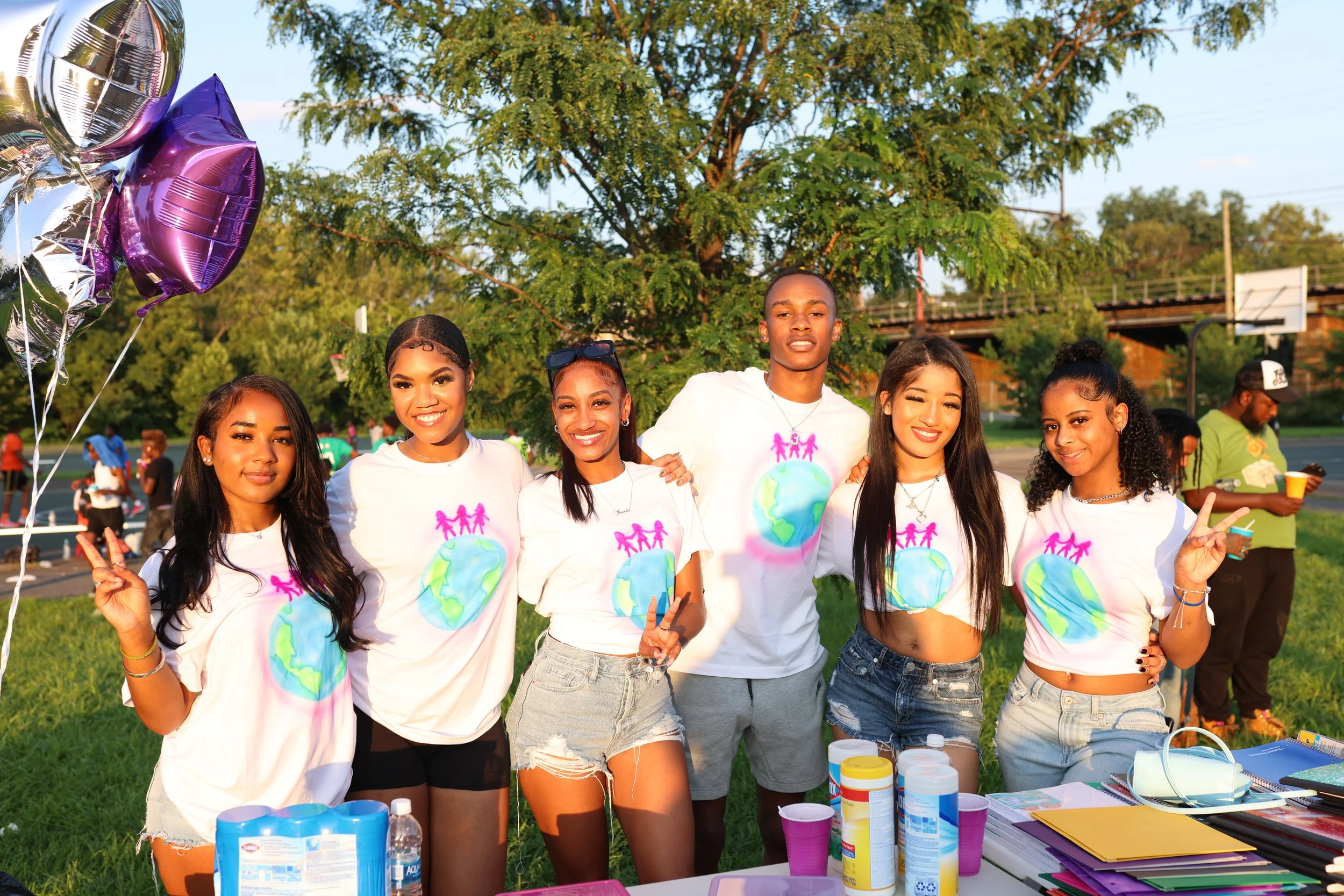 Six young women smiling and standing together outdoors near a table with arts and crafts supplies, wearing white T-shirts with a design of the Earth and pink human figures holding hands, under a large tree with green foliage in the background.