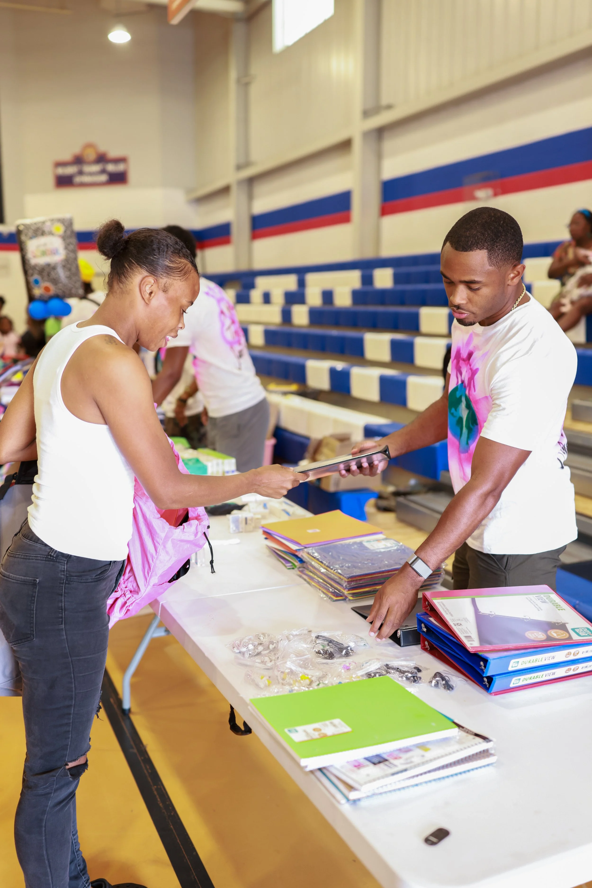 People at a table in a gymnasium with stacks of folders, notebooks, and miscellaneous items, engaging in an activity or discussion.